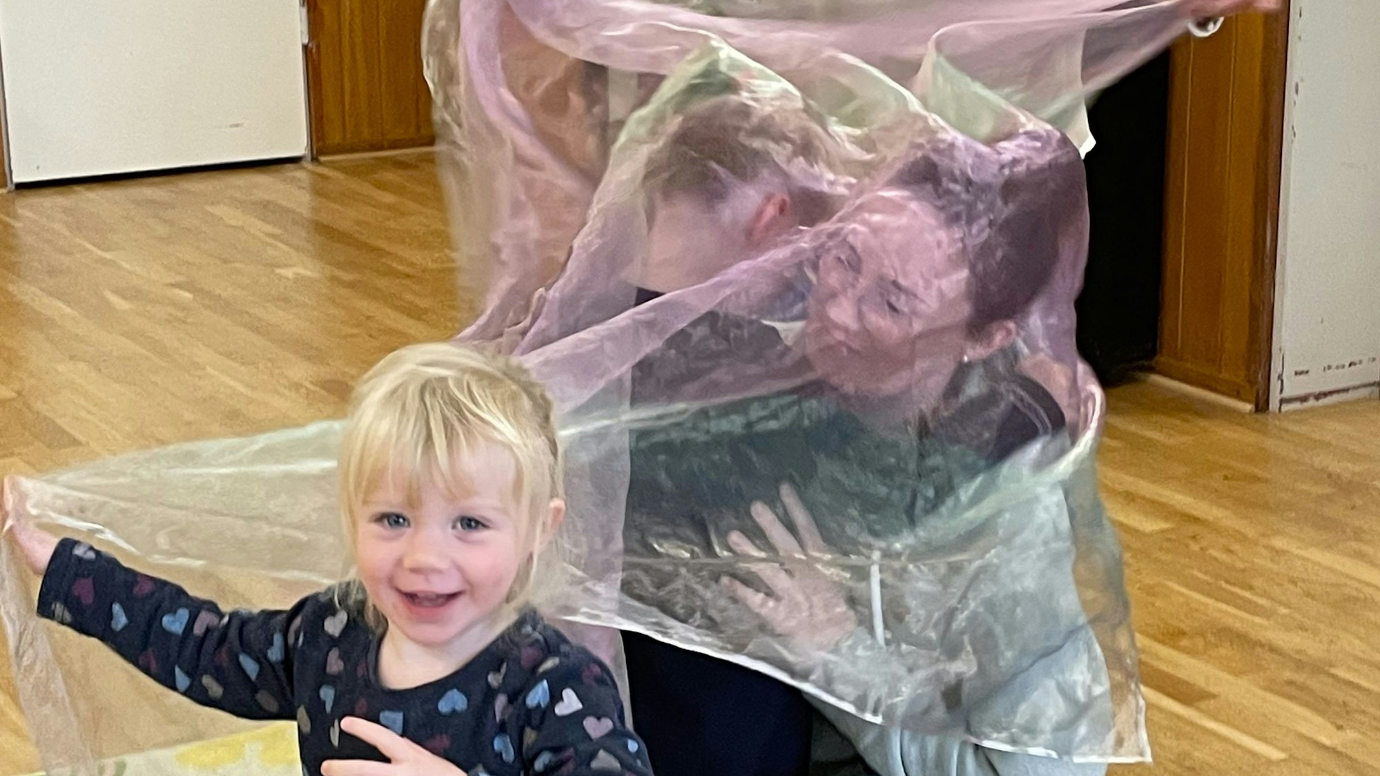 Ein kleines, blondes Mädchen lacht, hinter ihr sind ein anderes Mädchen und eine Frau komplett in ein Seidentuch gehüllt.