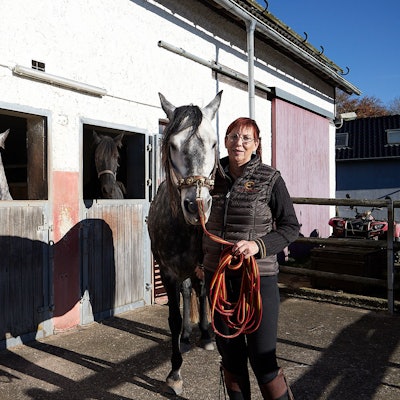 Eine Frau steht nah bei einem Pferd. Sie befinden sich neben einem Pferdestall, aus dem zwei weitere Tiere die Köpfe heraushalten.