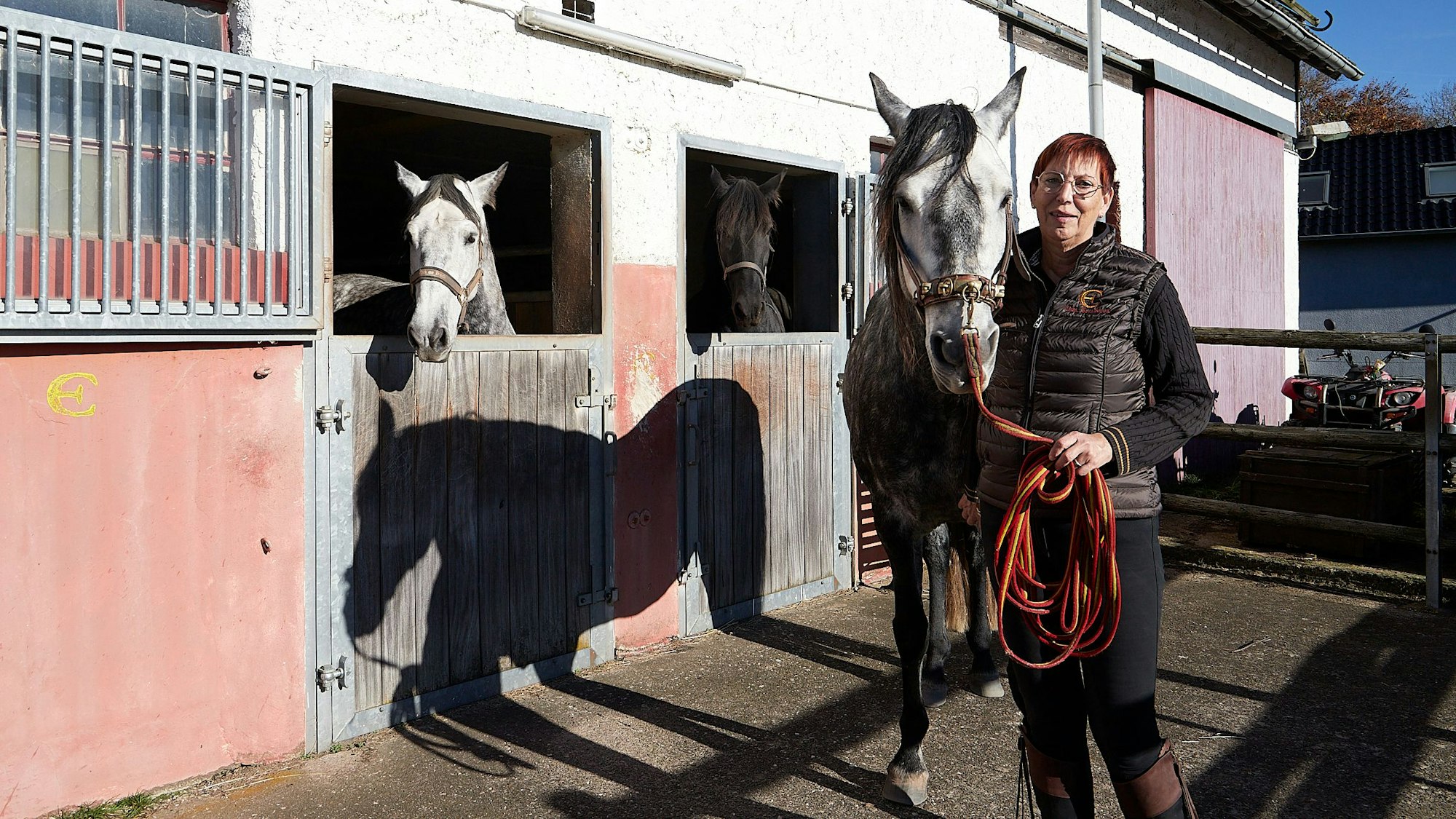 Eine Frau steht nah bei einem Pferd. Sie befinden sich neben einem Pferdestall, aus dem zwei weitere Tiere die Köpfe heraushalten.