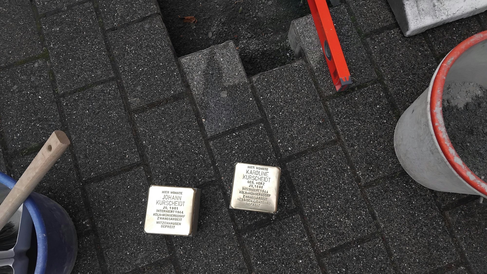 Zwei Stolpersteine mit den Namen Karoline Kurscheidt und Johann Kurscheidt liegen kurz vor ihrer Verlegung auf dem Bordstein, daneben sind Eimer und Werkzeuge zu sehen.