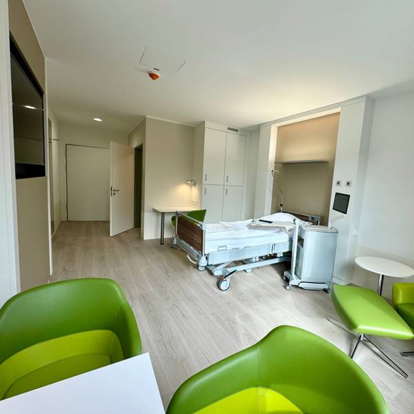 Auf dem Bild ist eines der Zimmer zu sehen, mit einem Krankenhausbett und grünen Sesseln.