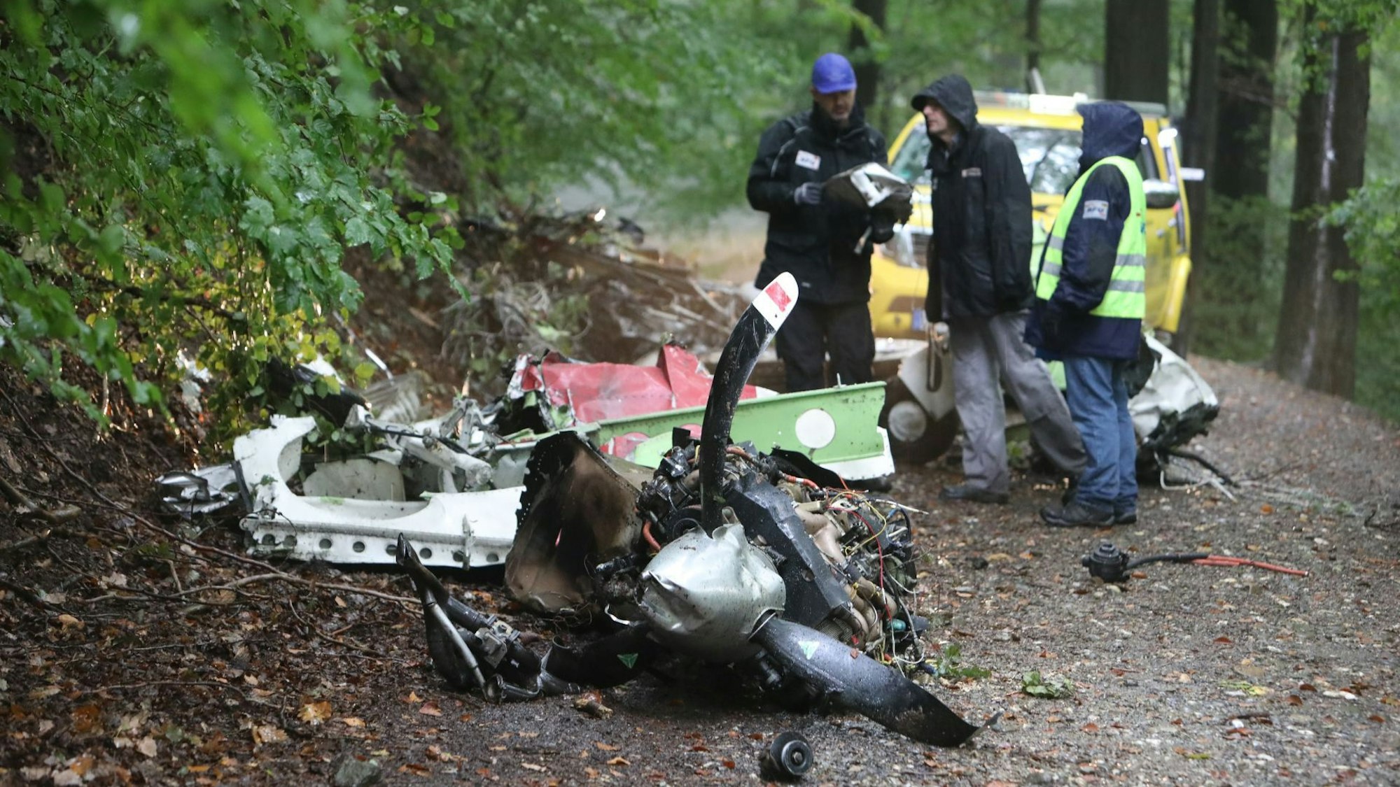 Trümmerteile liegen auf einem Waldweg, dahinter stehen drei Männer.