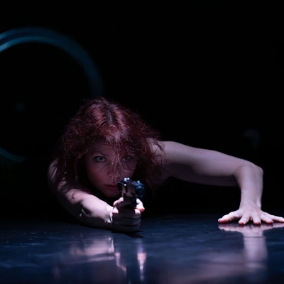 Eine Frau liegt mit ausgestreckter Hand auf dem Boden und zielt mit einer Pistole.