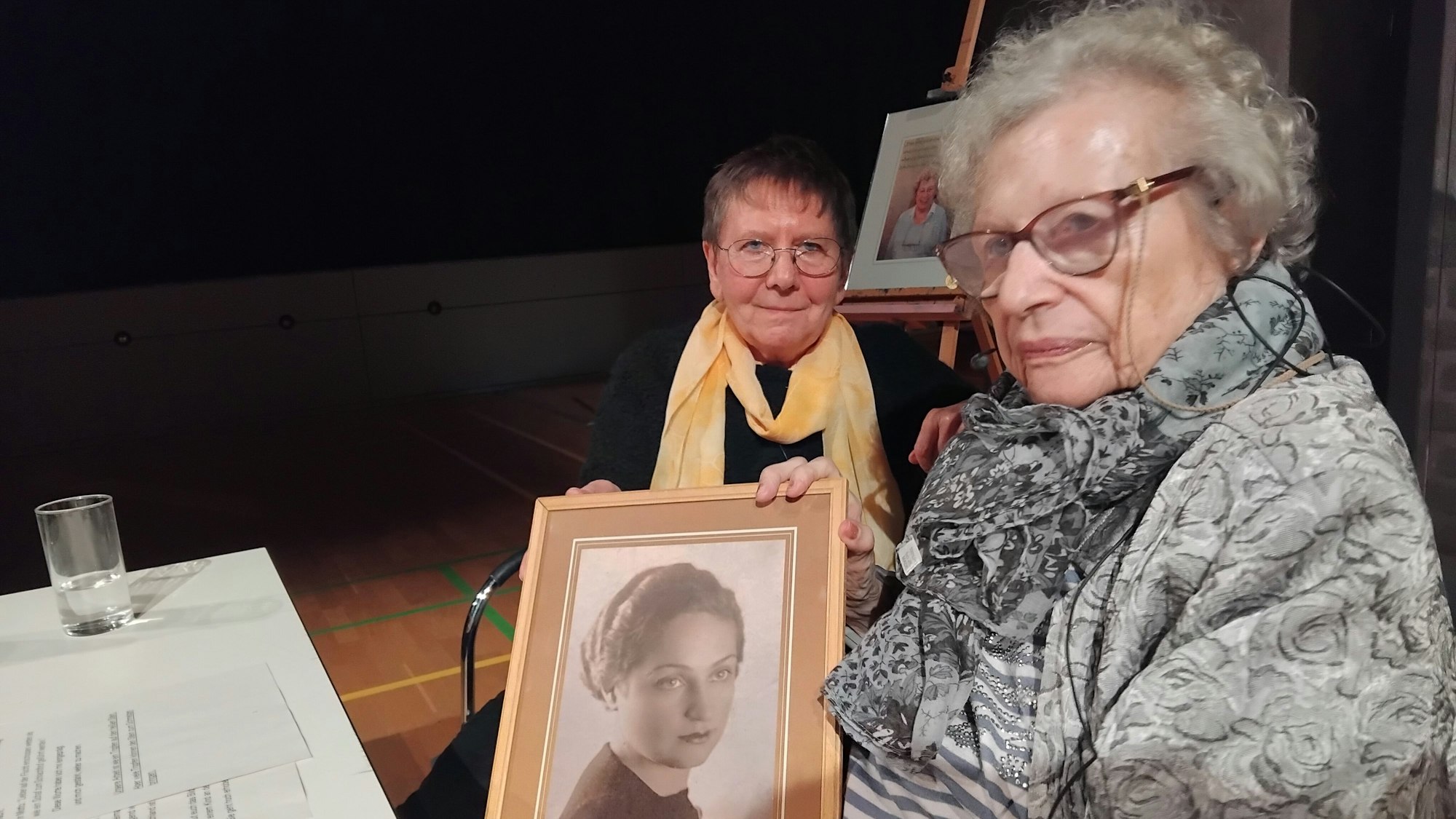 Zwei Frauen sitzen an einem Tisch, Tamar Dreifuss hält ein gerahmtes Schwarz-Weiß-Bild einer jungen Frau.