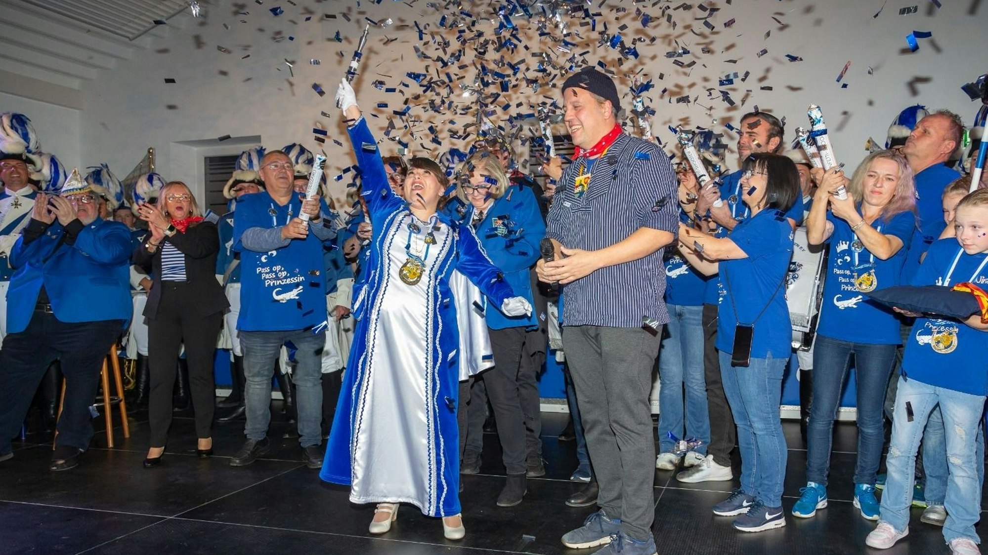 Anna Marian steht im blau-weißen Prinzessinnen-Orient auf einer Bühne und reckt jubelnd ein Zepter in die Höhe. Neben ihr steht Sacha Reichelt in grauer Hose und blau-weiß gestreiften Hemd. Im Hintergrund stehen viele Menschen in blauen T-Shirts, die Konfettikanonen in der Hand halten. Über alle regnet es silbernes und blaues Konfetti.