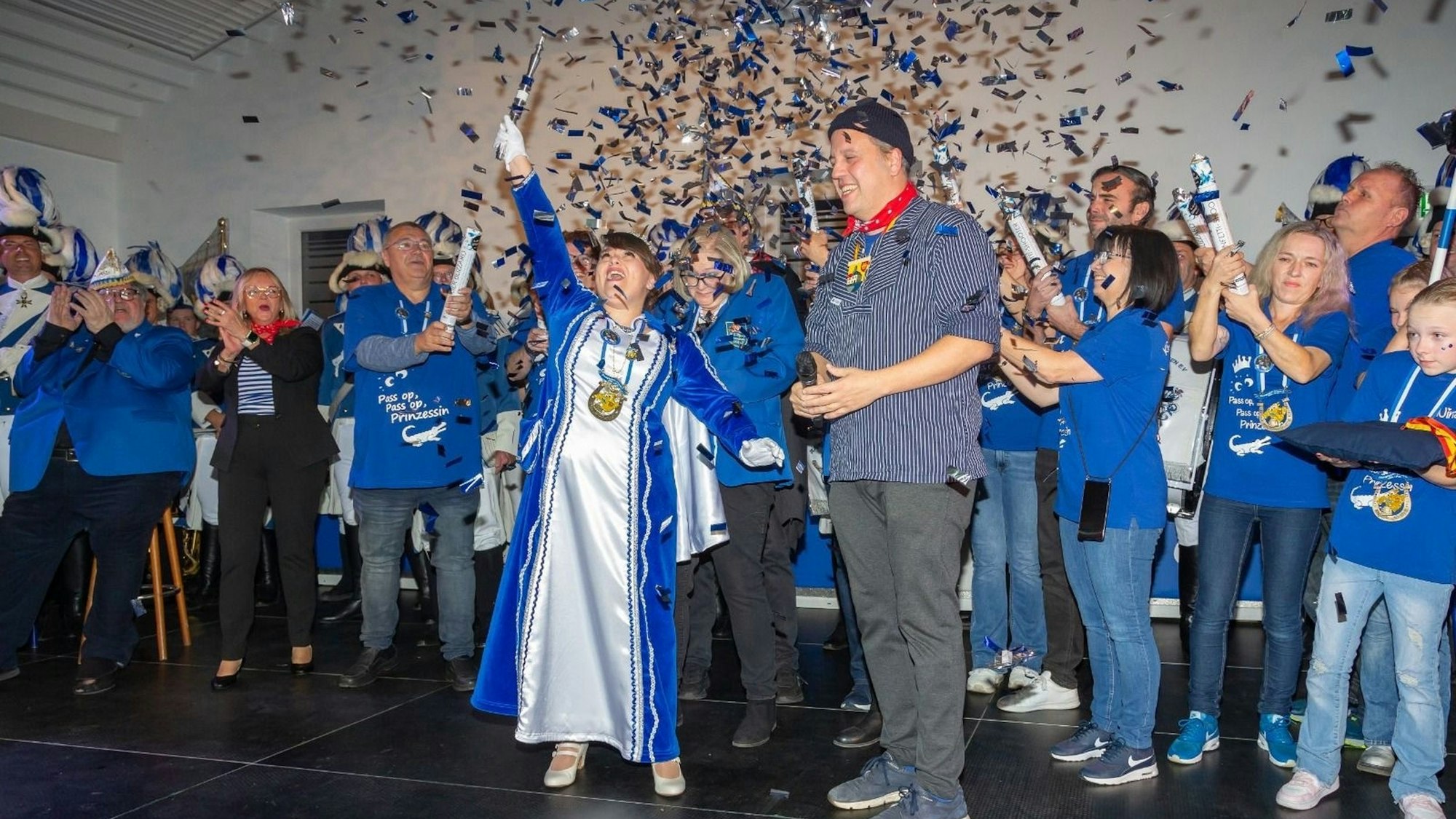 Anna Marian steht im blau-weißen Prinzessinnen-Orient auf einer Bühne und reckt jubelnd ein Zepter in die Höhe. Neben ihr steht Sacha Reichelt in grauer Hose und blau-weiß gestreiften Hemd. Im Hintergrund stehen viele Menschen in blauen T-Shirts, die Konfettikanonen in der Hand halten. Über alle regnet es silbernes und blaues Konfetti.