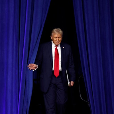 Donald Trump schreitet durch einen blauen Vorhang auf eine Bühne.