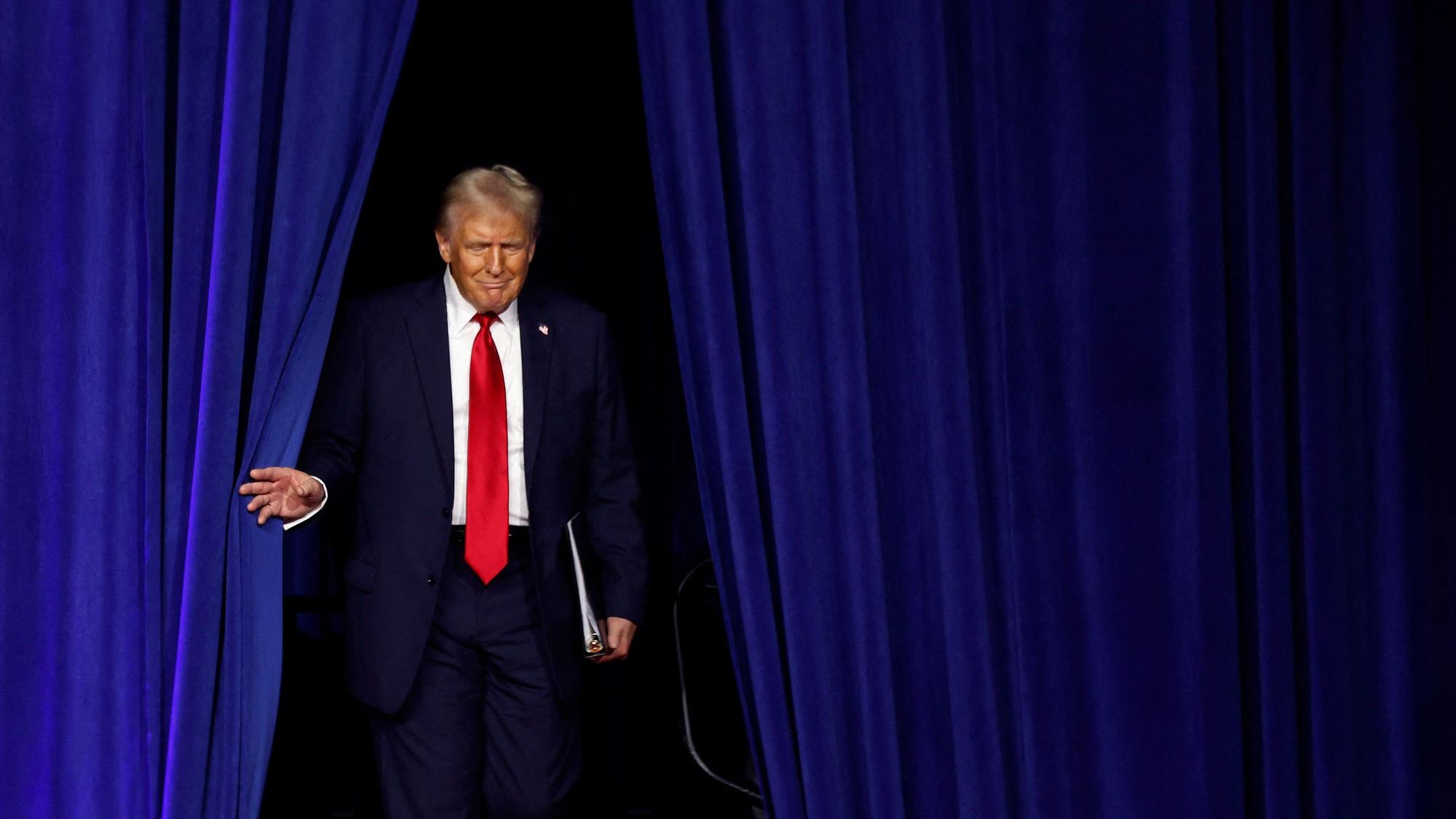 Donald Trump schreitet durch einen blauen Vorhang auf eine Bühne.