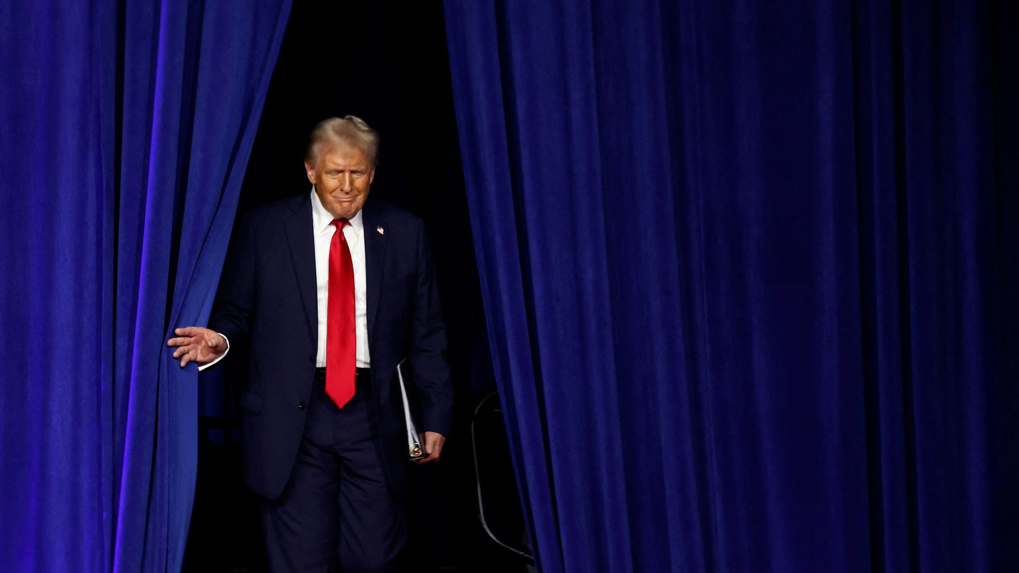 Donald Trump schreitet durch einen blauen Vorhang auf eine Bühne.