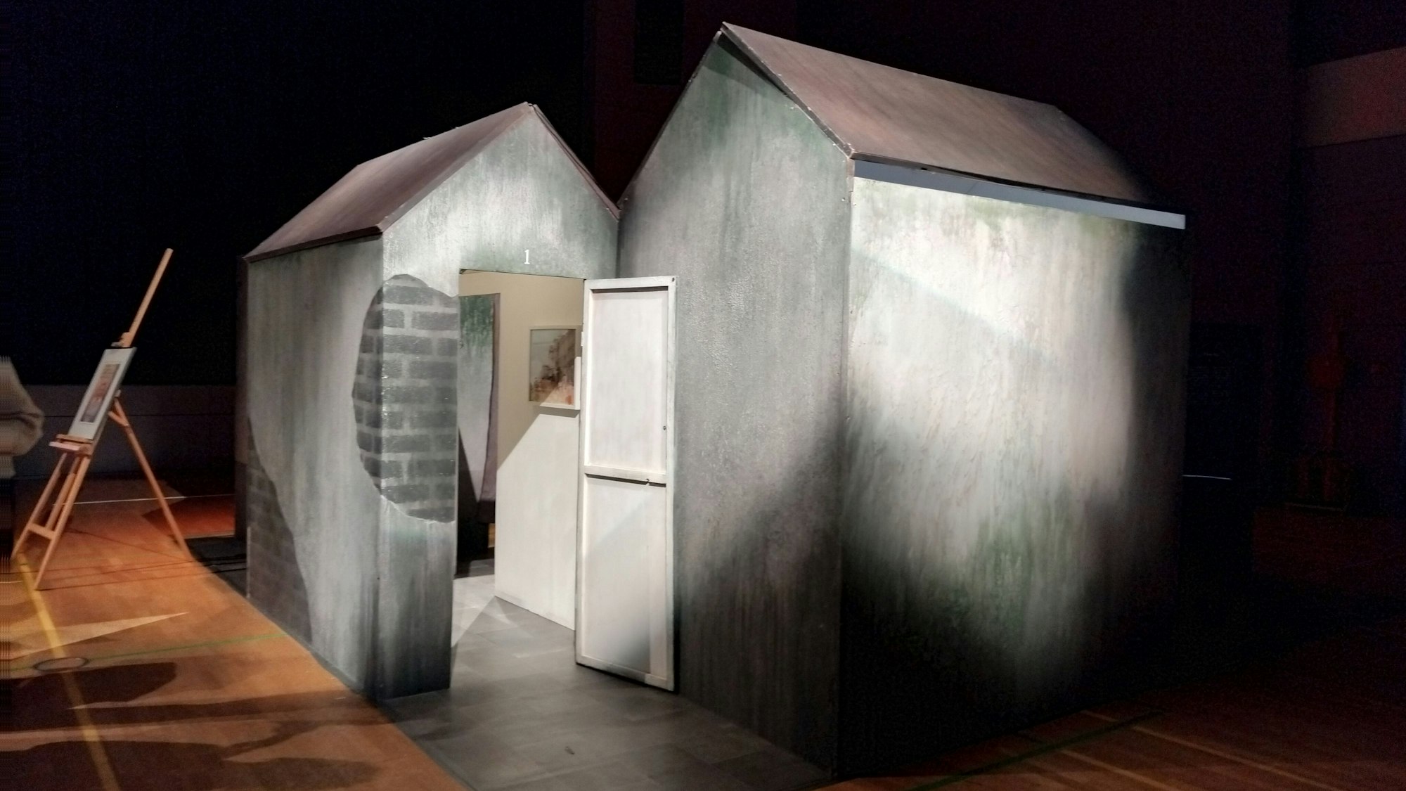 Eine Installation in Form eines grauen Hauses steht beleuchtet in einer Turnhalle.