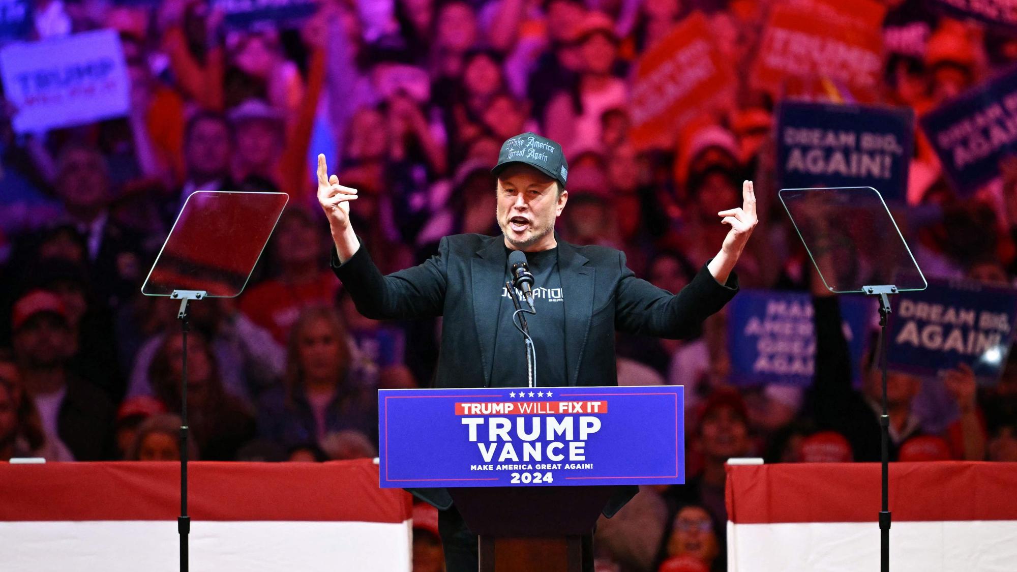 Elon Musk hält eine Rede an einem Pult mit der Aufschrift "Trump, Vance".