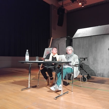 Zwei Frauen sitzen an einem Tisch in einer Turnhalle, im Hintergrund sieht man eine Installation in Form eines Hauses.