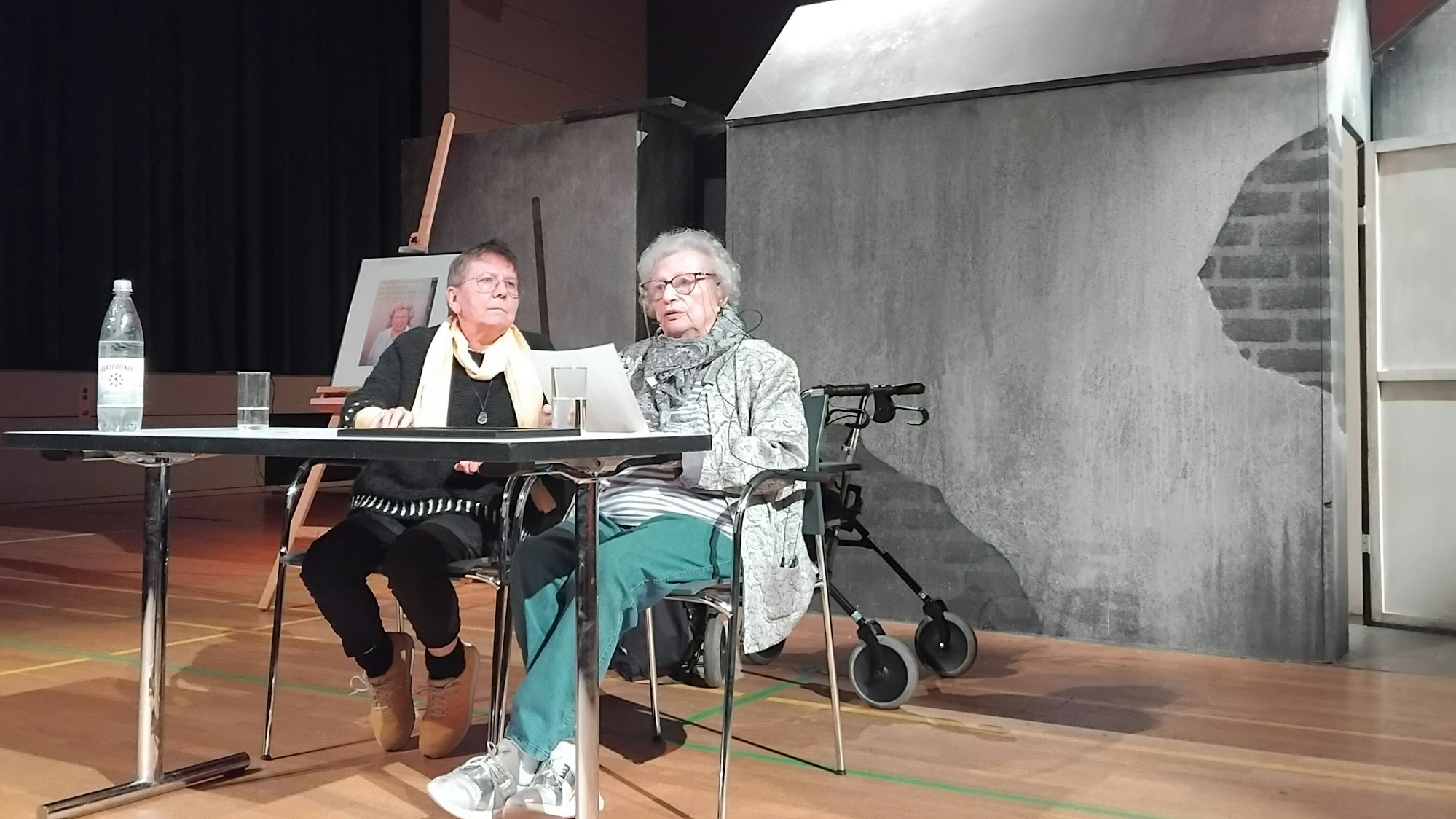 Zwei Frauen sitzen an einem Tisch in einer Turnhalle, im Hintergrund sieht man eine Installation in Form eines Hauses.