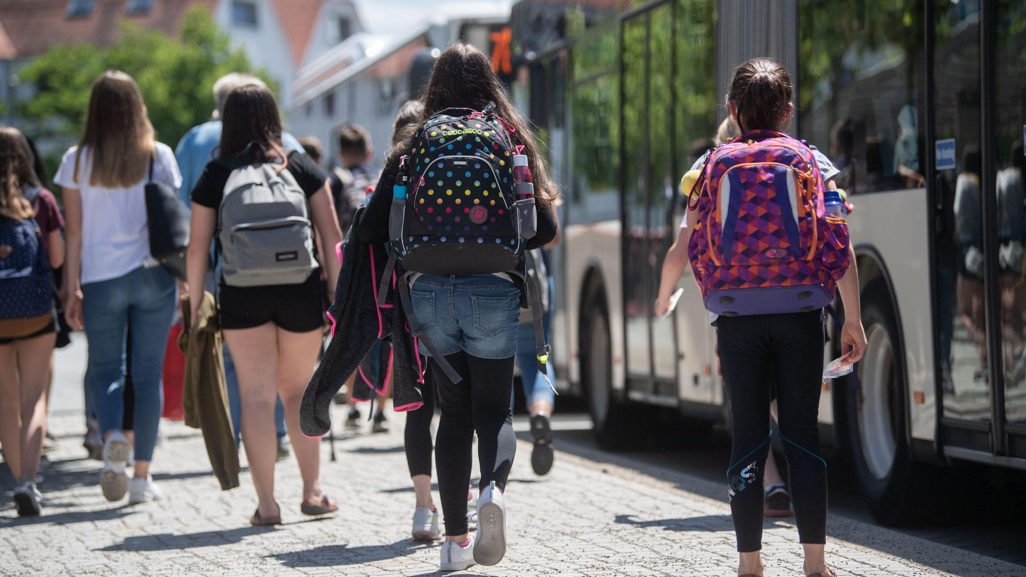 Schülerinnen, manche mit Schulranzen auf dem Rücken, gehen an einer Bushaltestelle zu stehenden Bussen. Die Mädchen sind von hinten zu sehen.