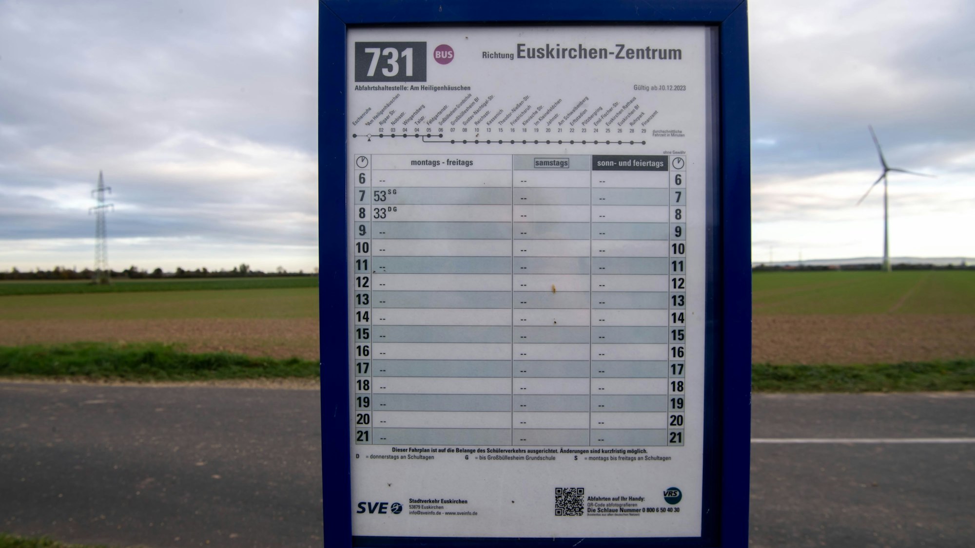 Ein Fahrplan der Linie 731 Richtung Euskirchen-Zentrum ist mittig im Bild abgelichtet, im Hintergrund Felder mitsamt Strommast und Windkraftrad.