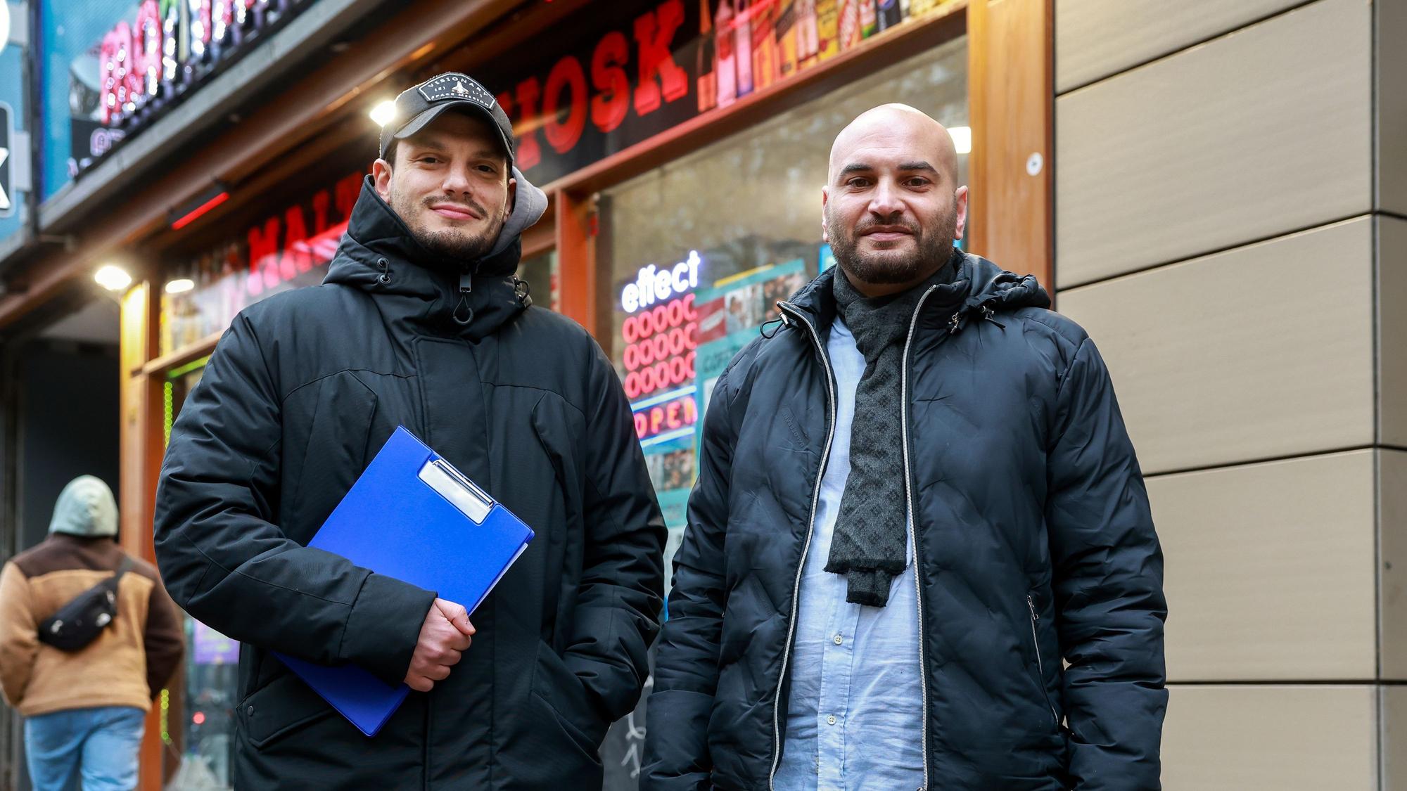 Ibrahim Cuma und Malik Dine stehen mit einem Klemmbrett vor einem Kiosk