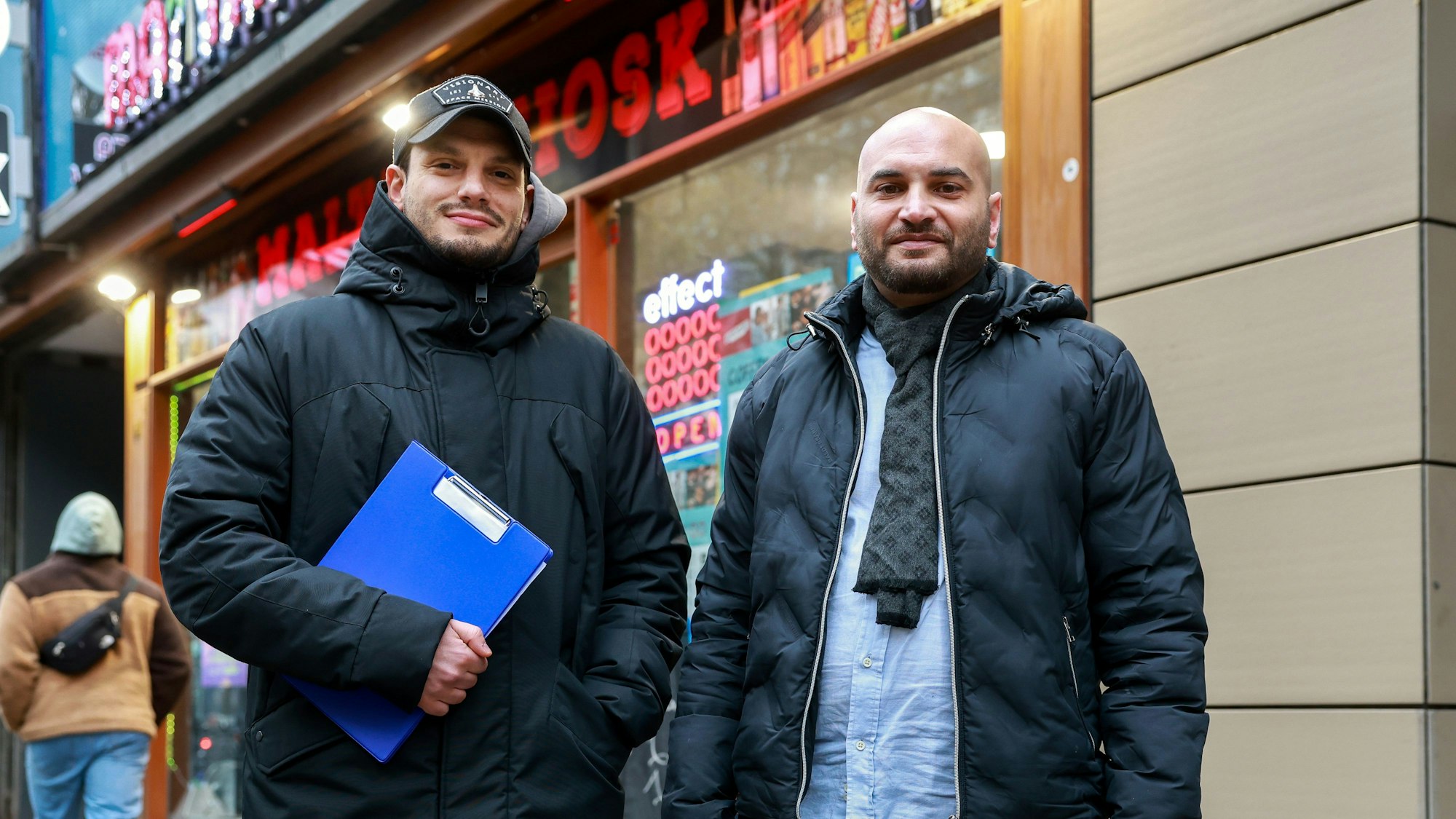Ibrahim Cuma und Malik Dine stehen mit einem Klemmbrett vor einem Kiosk