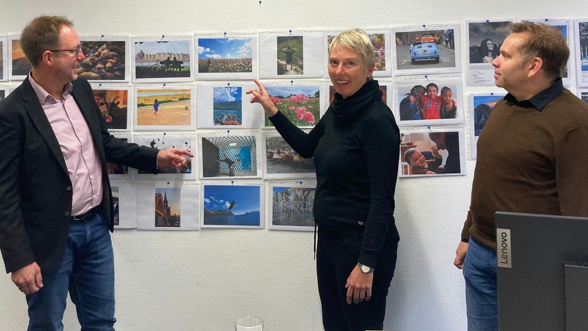Zu sehen sind zwei Männer und eine Frau vor einer Wand mit Fotos.
