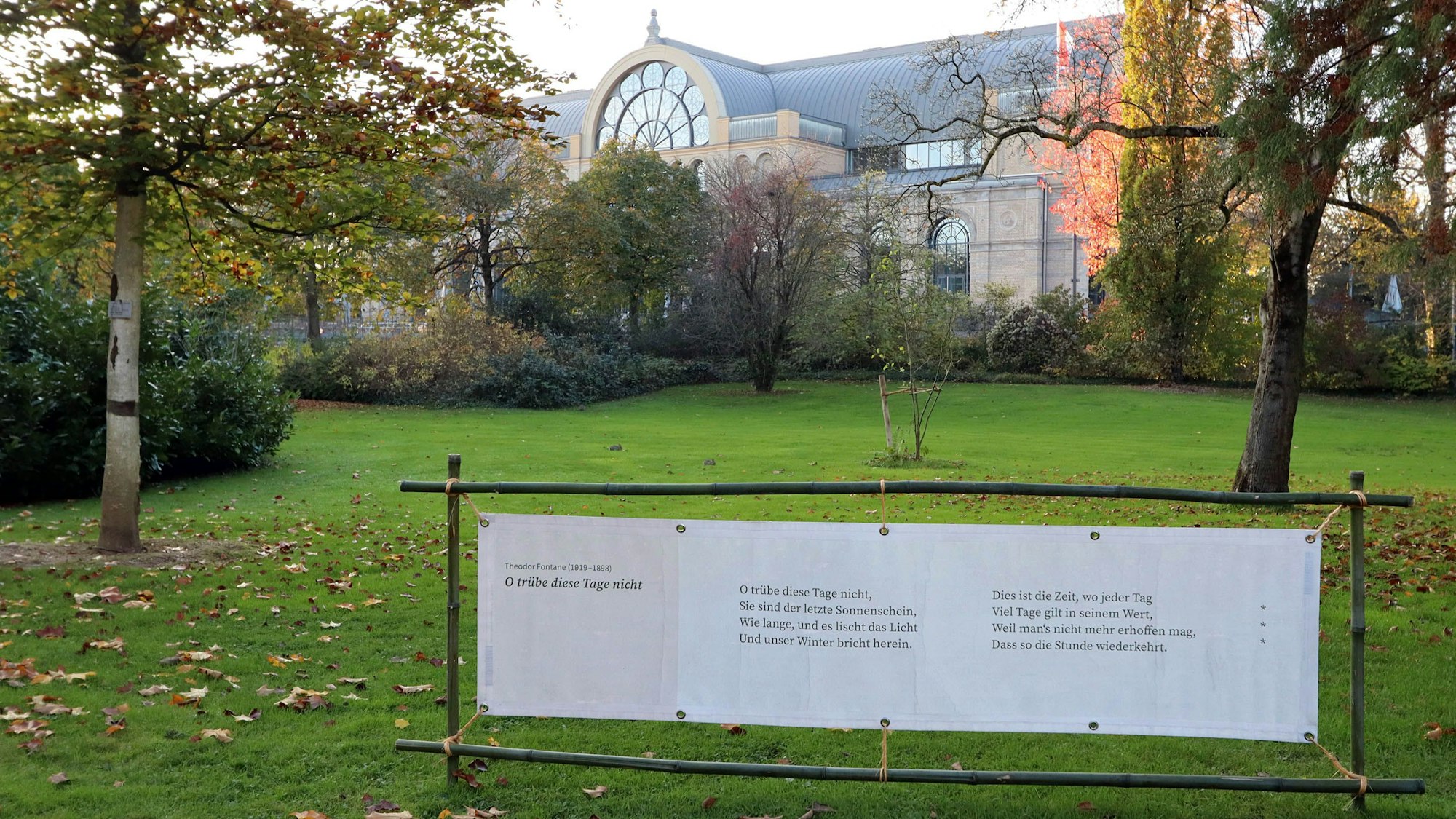 Auf einer Wiese steht ein breiter Aufsteller mit einem Gedicht darauf, im Hintergrund ist das Gebäude der Flora zu sehen.