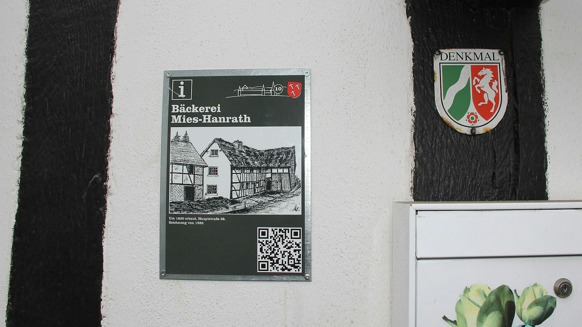 An einem Haus ist eine Tafel angebracht, die eine Zeichnung und einen QR-Code zeigt.