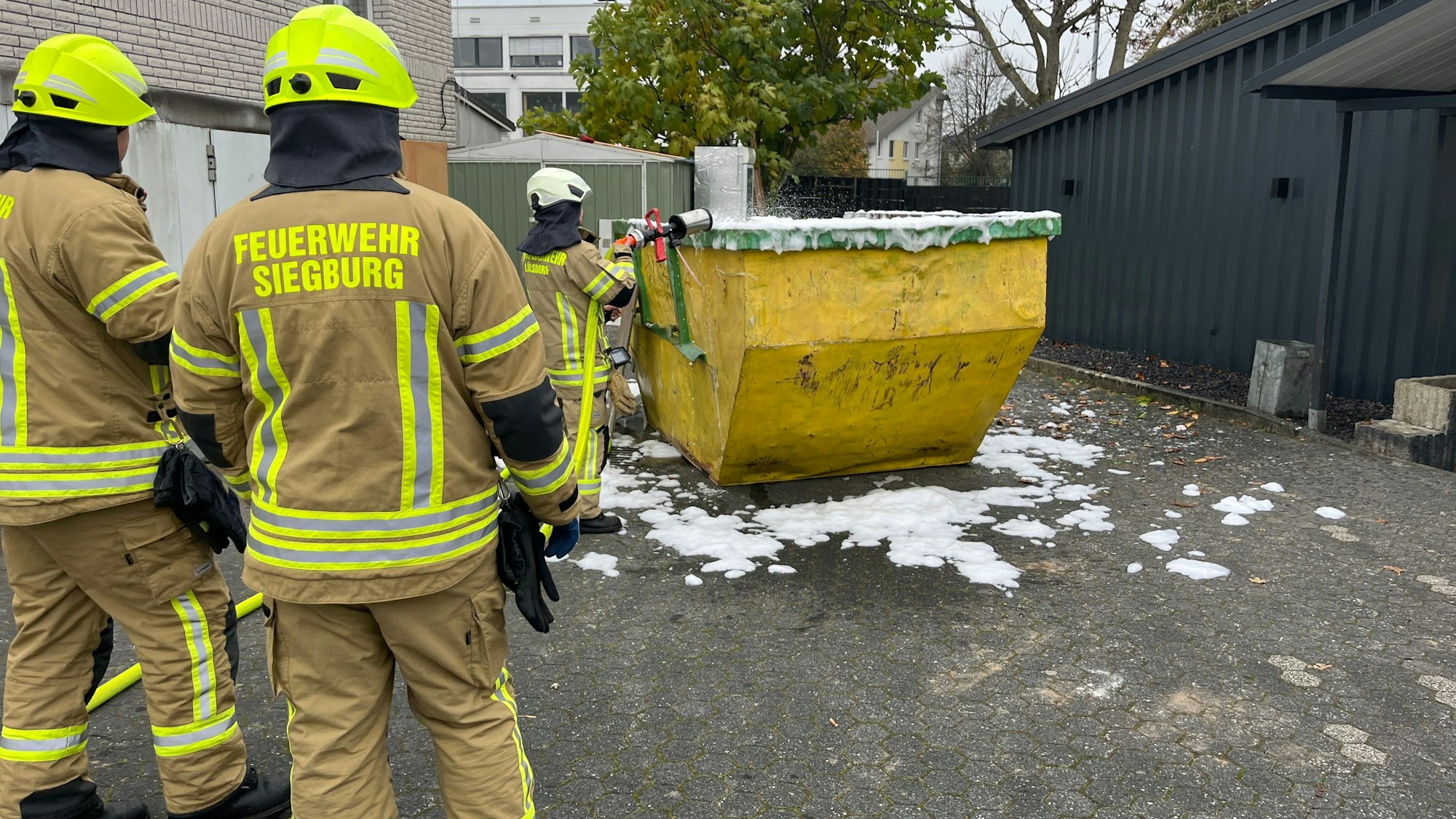 Drei Feuerwehrleute stehen vor einem Container, der mit Löschschaum geflutet wurde.