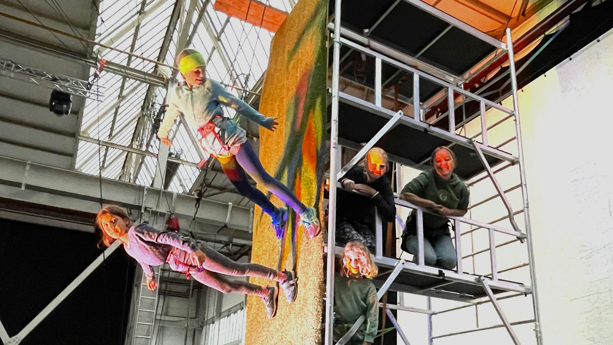 Bei den Proben des Kölner Flugtheaters laufen zwei Mädchen, die mit Seilen gesichert sind, senkrecht an einer Wand entlang, daneben stehen drei andere Mädchen auf einem Gerüst.
