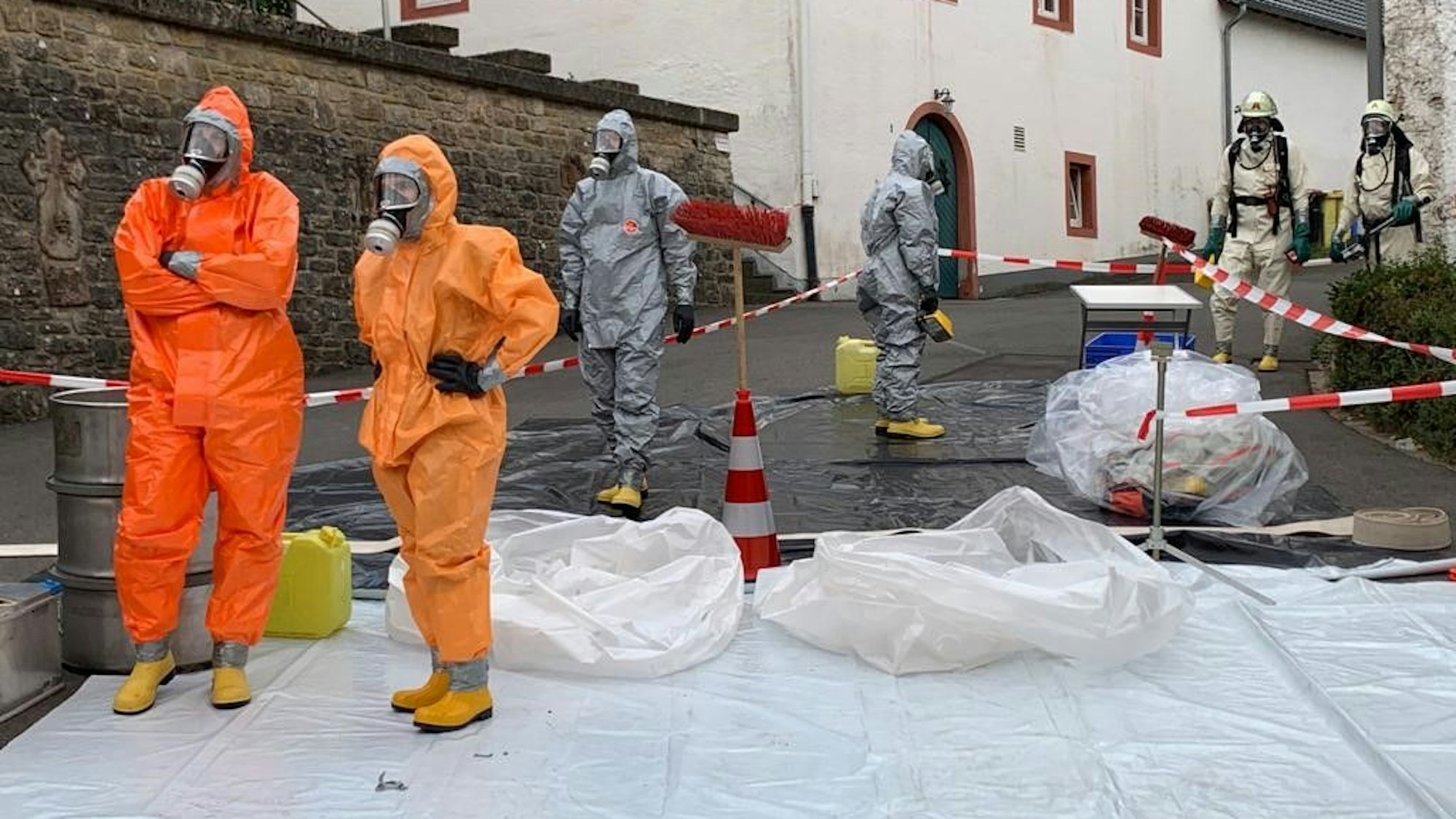 Einsatzkräfte in orangefarbenen und grauen Schutzanzügen stehen auf einer weißen Plane vor einem Haus.