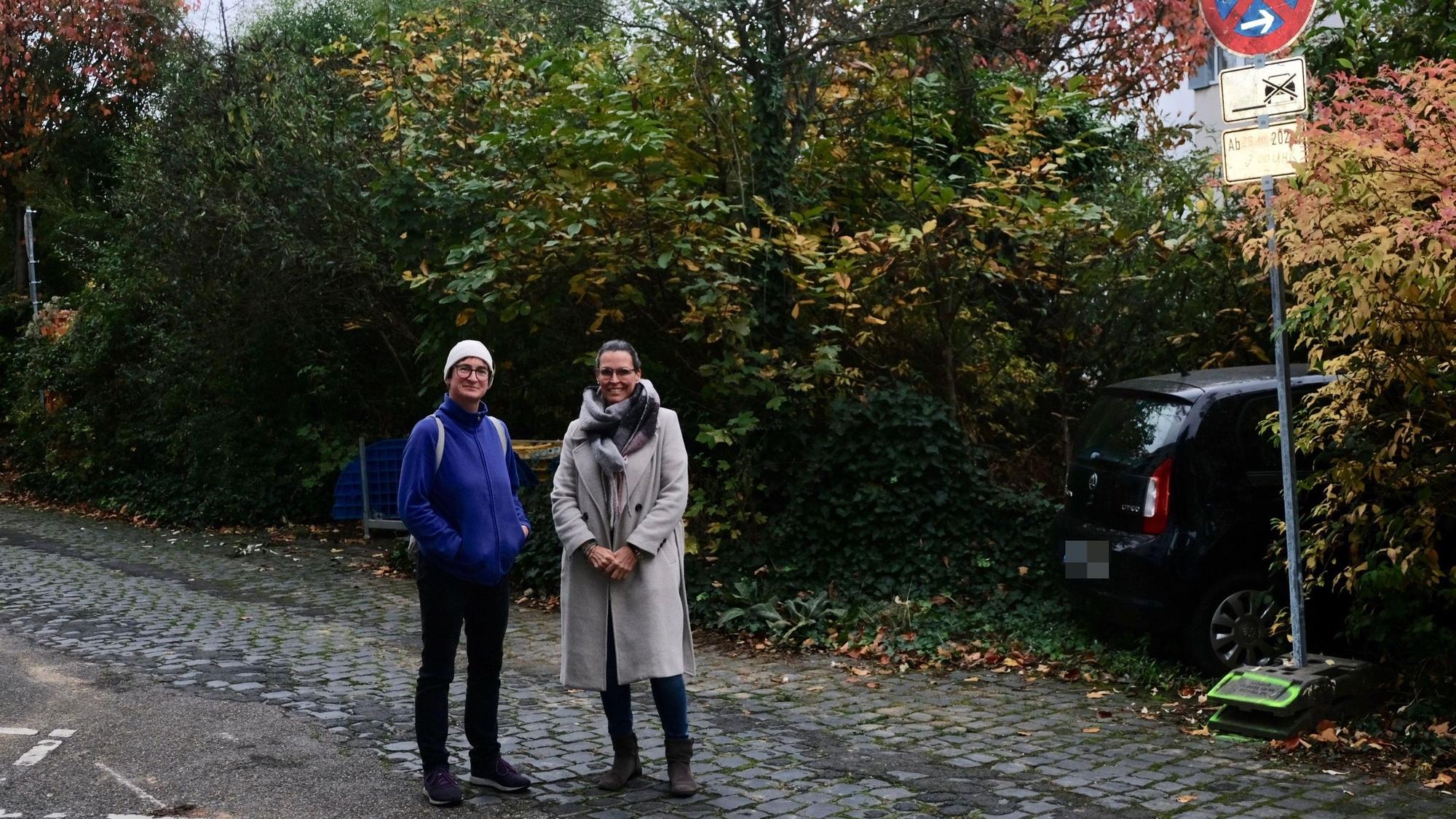 Zwei Frauen stehen auf einer Straße, hinter ihnen ist üppig wucherndes Grün zu sehen.