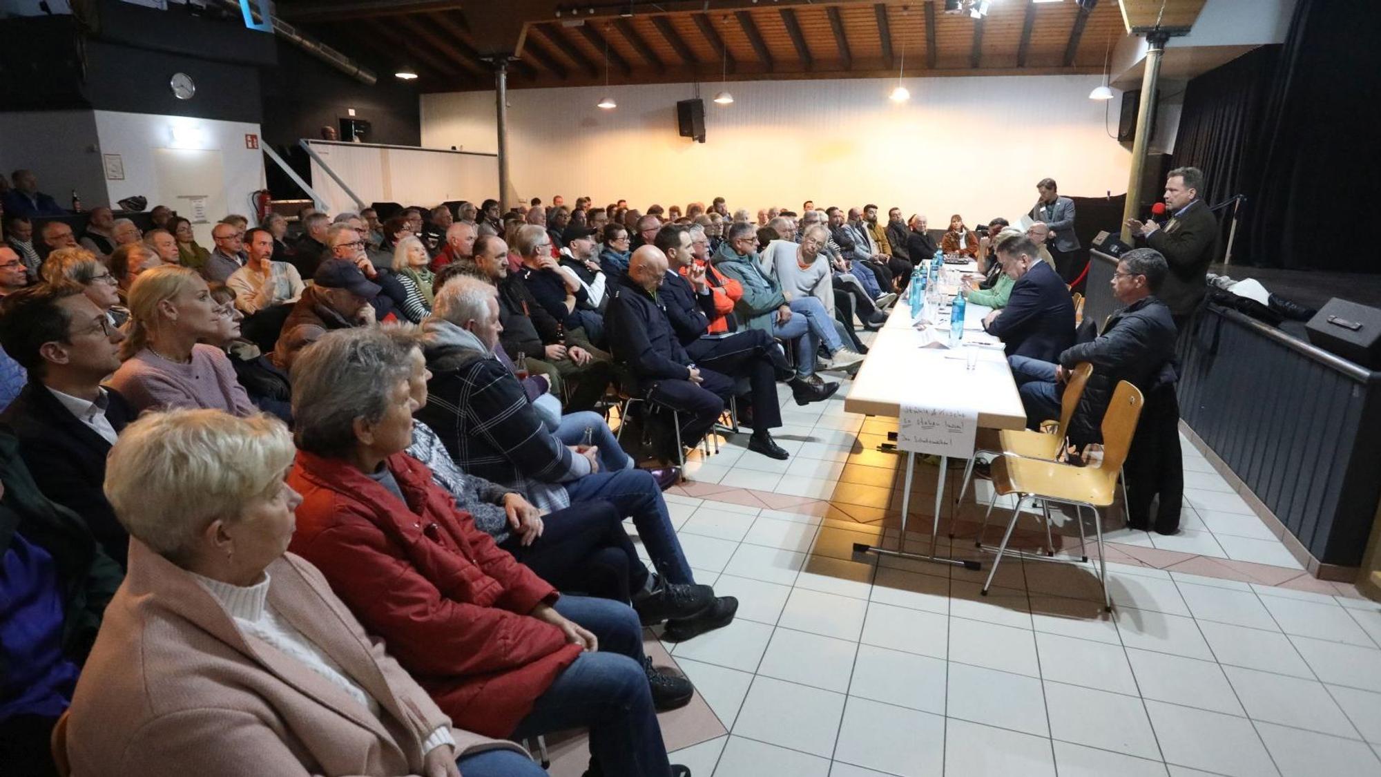 Zahlreiche Menschen sitzen in einem Saal und hören einem Mann mit Mikrofon zu.