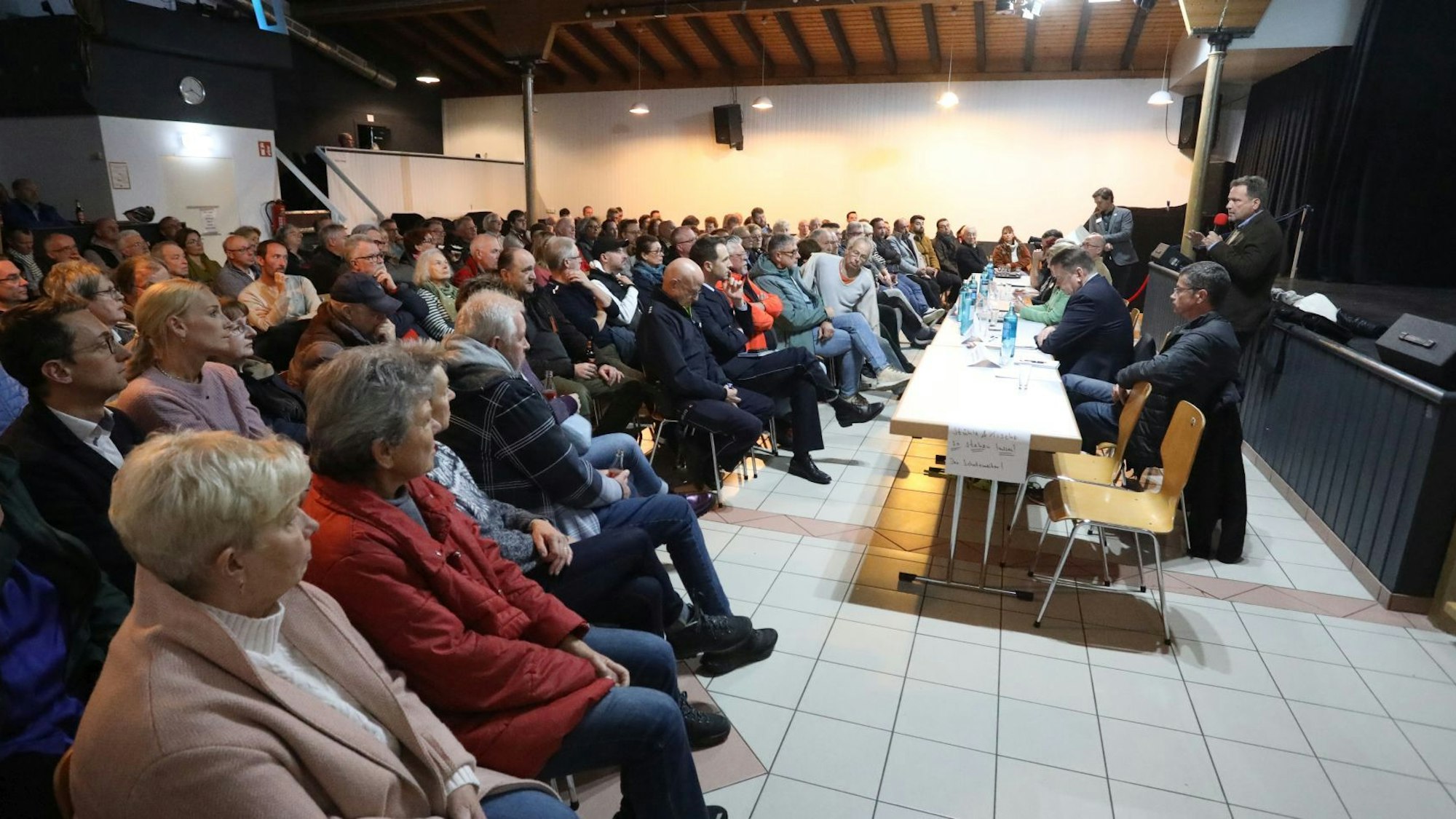 Zahlreiche Menschen sitzen in einem Saal und hören einem Mann mit Mikrofon zu.
