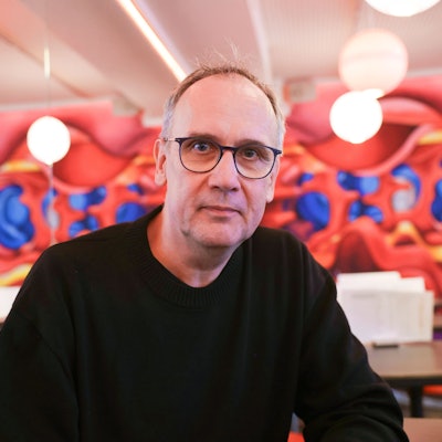 Volker Kutscher sitzt in einem Café vor einer bunten Wand. Er trägt einen schwarzen Pullover.