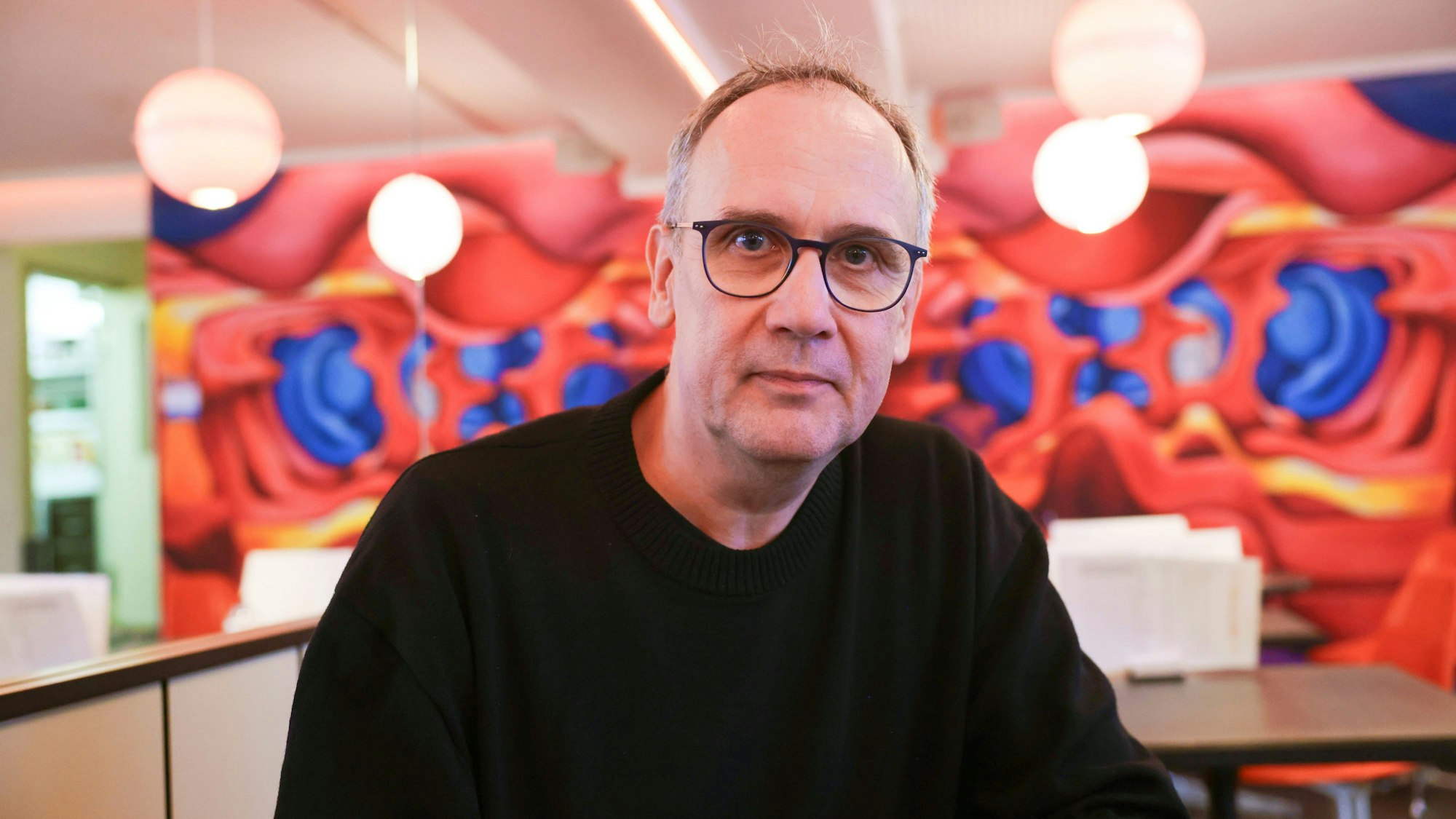 Volker Kutscher sitzt in einem Café vor einer bunten Wand. Er trägt einen schwarzen Pullover.