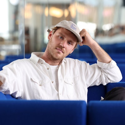 Zu sehen ist der in weiß gekleidete Musiker Jules Ahoi, der auf einem blauen Sessel sitzt und eine beige Kappe trägt.