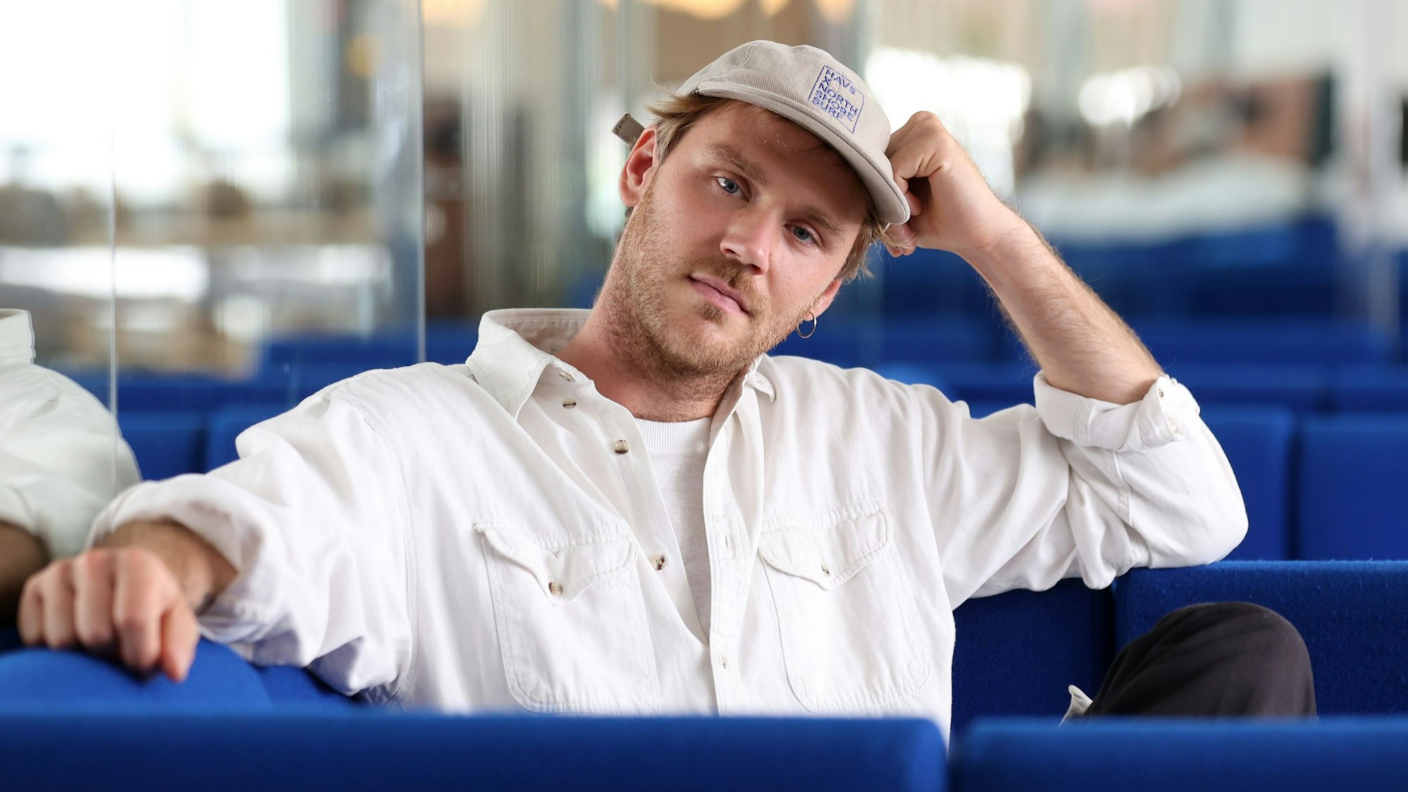 Zu sehen ist der in weiß gekleidete Musiker Jules Ahoi, der auf einem blauen Sessel sitzt und eine beige Kappe trägt.