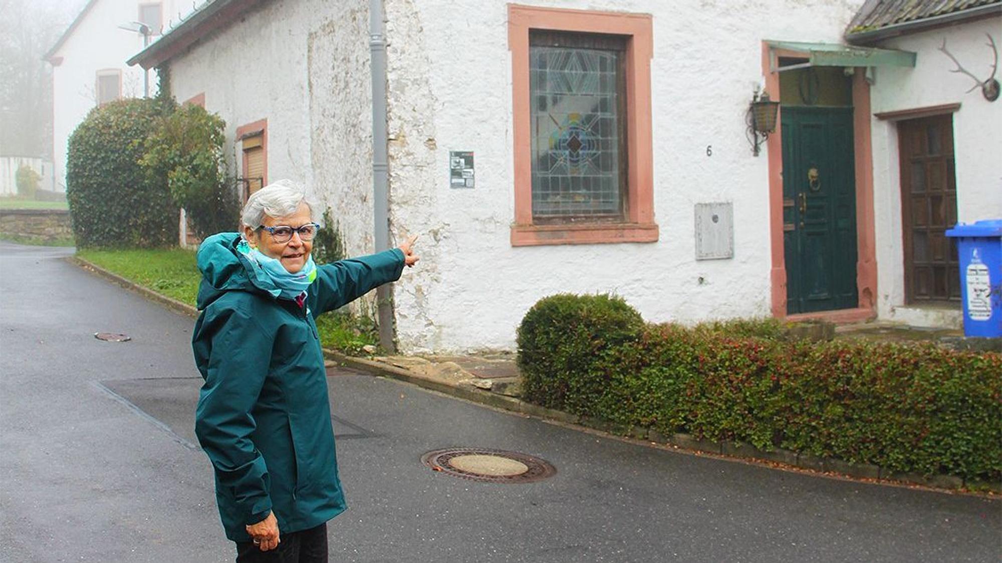 Eine Frau in einer blauen Regenjacke steht vor einem weißen Haus und zeigt auf die Fenster.