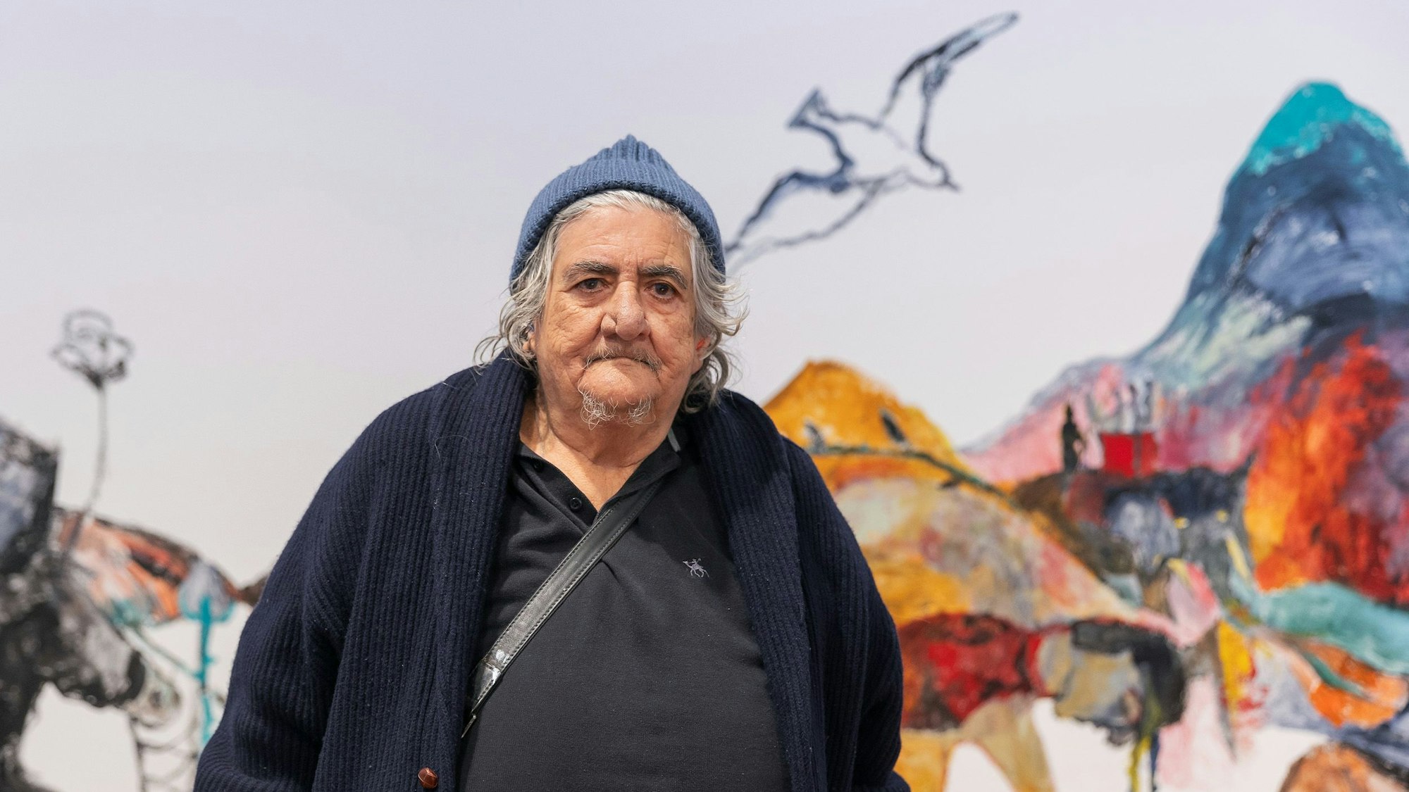 Anna Boghiguian trägt eine Mütze und eine blaue Strickjacke und schaut in die Kamera.