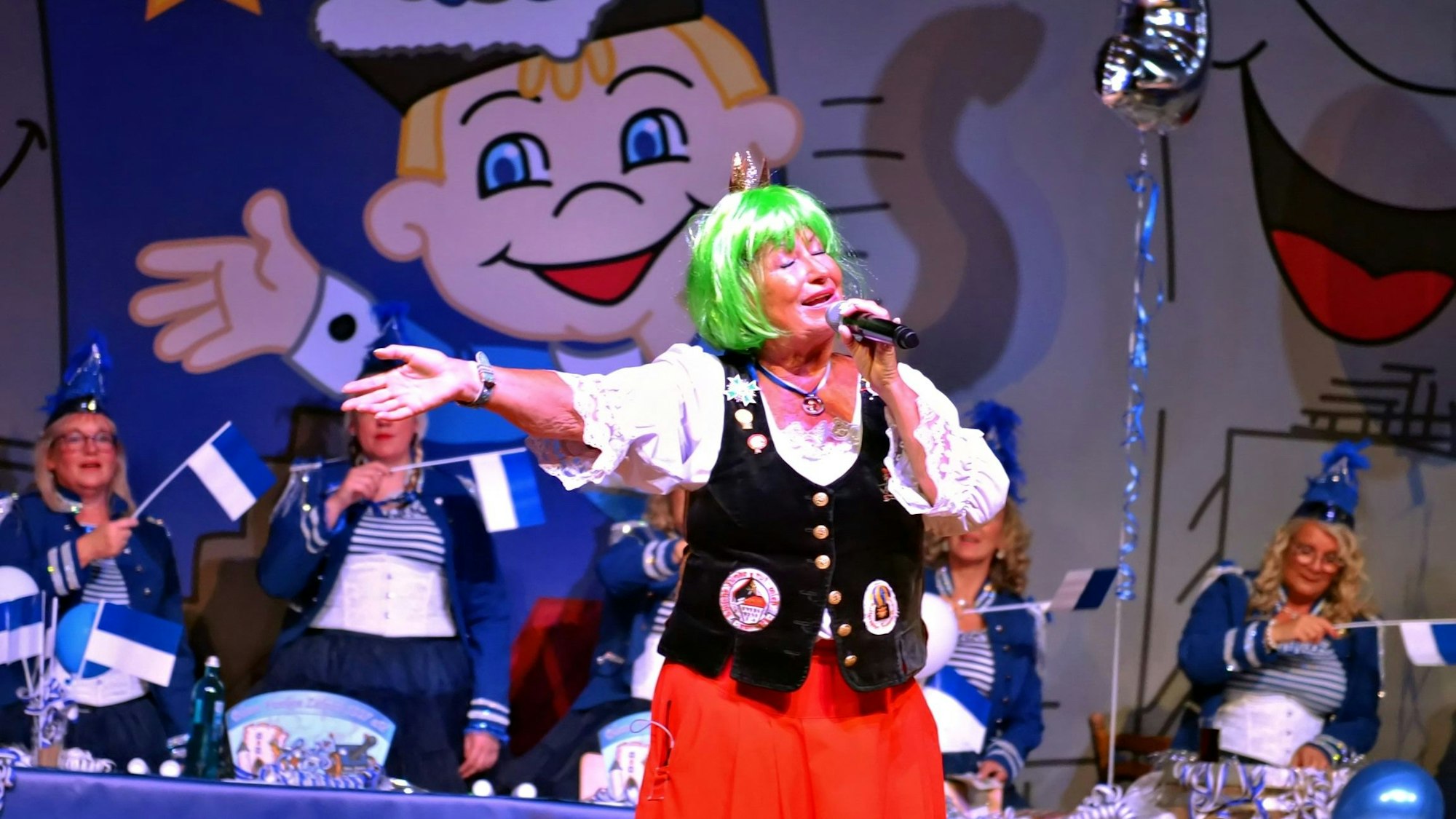 Eine Sängerin in Kostüm und mit grüner Perücke steht auf der Bühne, im Hintergrund schwenken Blaue Funken kleine Fahnen.