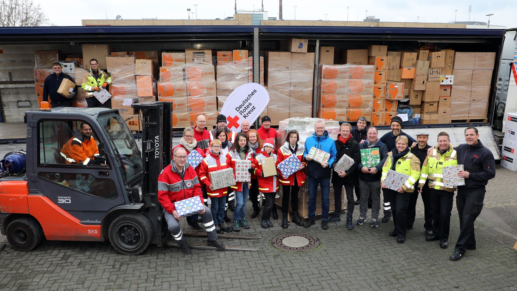 Menschen mit Paketen stehen neben einem Gabelstapler vor einem Sattelzugauflieger.
