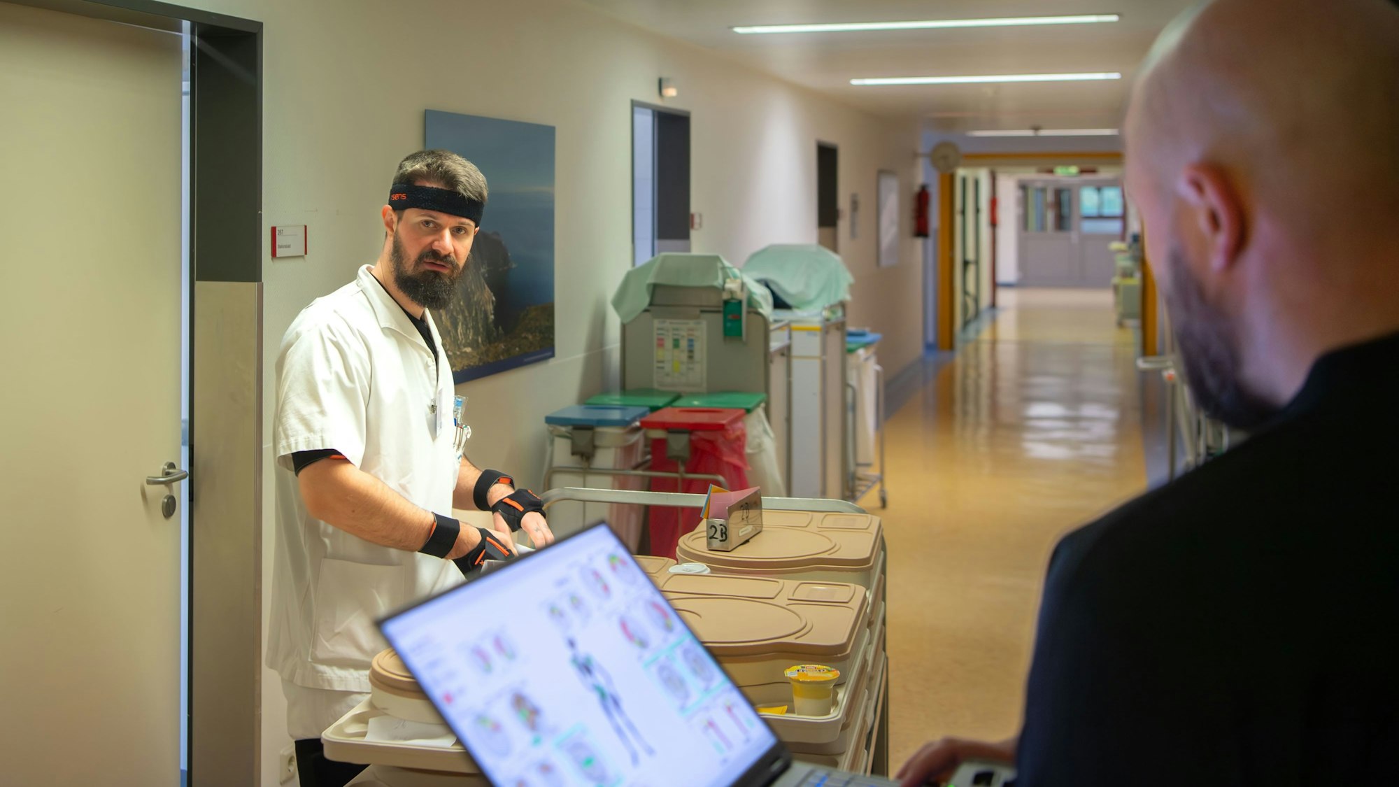 Ein Mitarbeiter in weißer Kleidung steht auf einem Krankenhausflur. An Kopf und Armen trägt er Sensoren. Ein anderer Mann an einem Laptop beobachtet ihn.