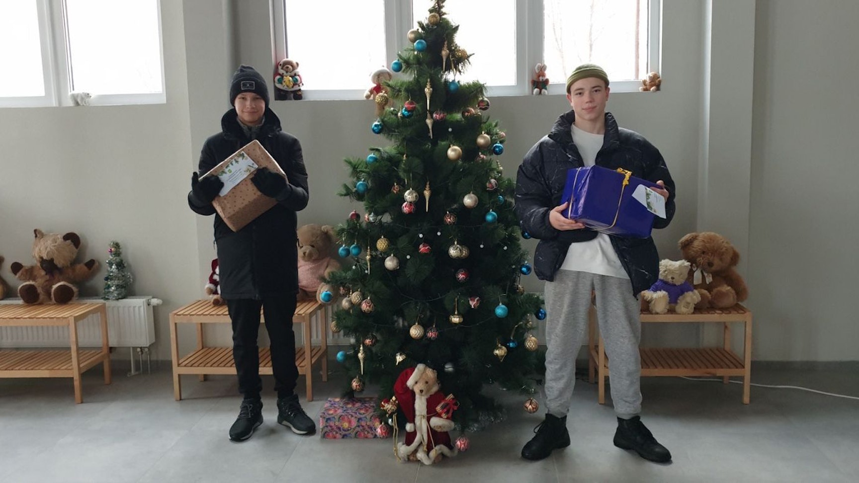 Kinder stehen mit zwei Paketen in Jacken und Mützen neben einem Weihnachtsbaum in einer Halle