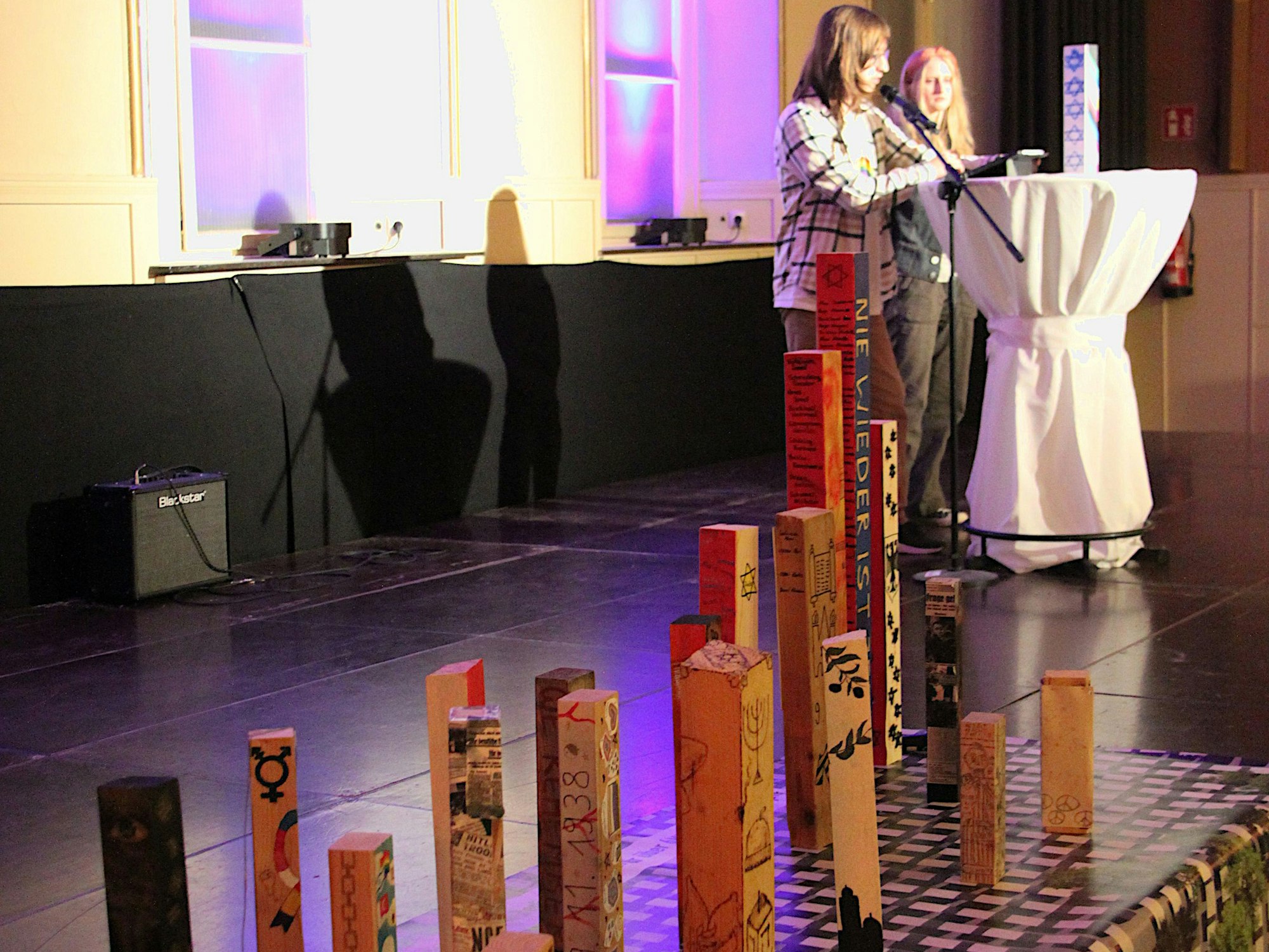 Zwei Schülerinnen an einem Stehtisch lesen etwas vor. Auf der Bühne neben ihnen stehen künstlerisch gestaltete Stelen.