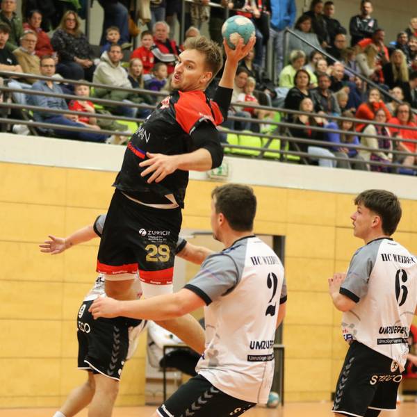 Handballer Robin Voiß steigt zum Sprungwurf hoch. Um ihn herum drei Gegenspieler, direkt vor ihm springt ein weiterer mit erhobenen Armen hoch. Im Hintergrund ist die Zuschauertribüne zu erkennen.