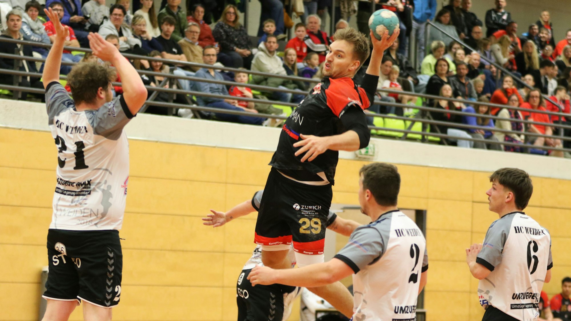 Handballer Robin Voiß steigt zum Sprungwurf hoch. Um ihn herum drei Gegenspieler, direkt vor ihm springt ein weiterer mit erhobenen Armen hoch. Im Hintergrund ist die Zuschauertribüne zu erkennen.