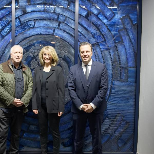 Eine Gruppe von drei Männern und einer Frau stehen vor einem wandgroßen, blauen Glaskunstwerk.