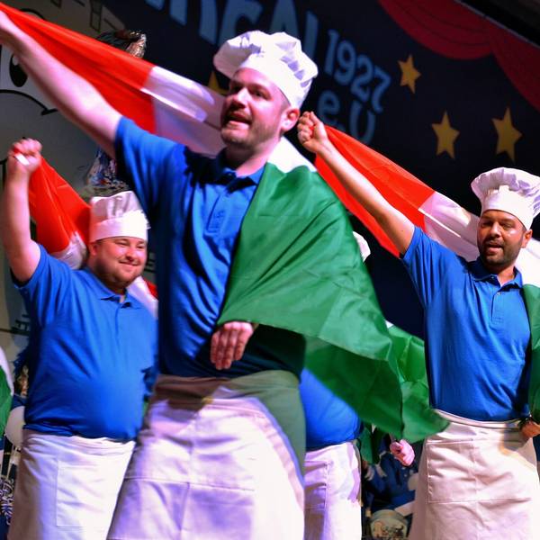 Als Köche verkleidete Männer stehen tanzend auf der Bühne und schwenken italienische Flaggen.