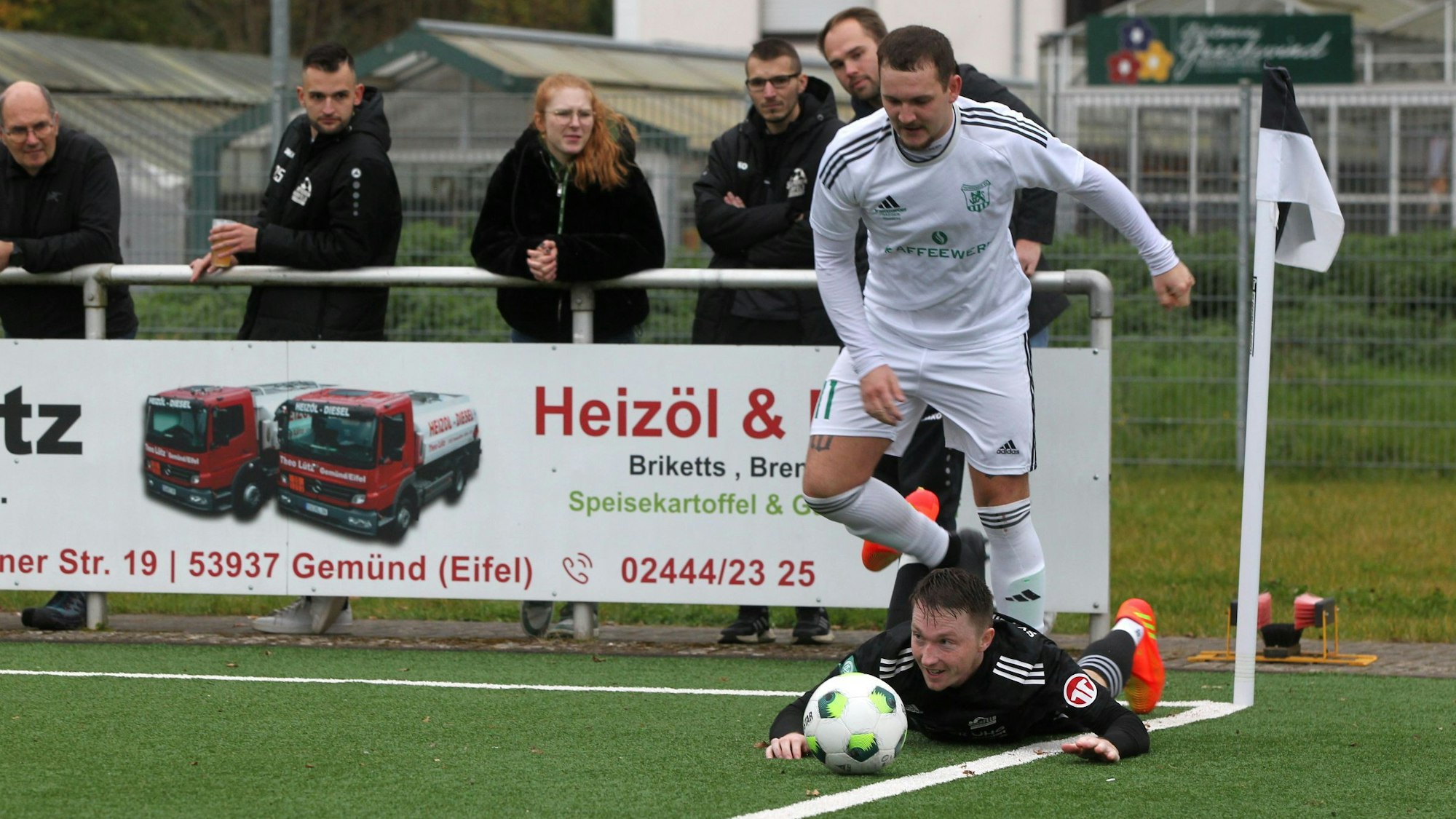 Ein Spieler des SV Nierfeld liegt nahe der Eckfahne am Boden. Der Gegner, der hinter ihm steht, hatte ihn gefoult. Zuschauer hinter der Bande beobachten das Geschehen.