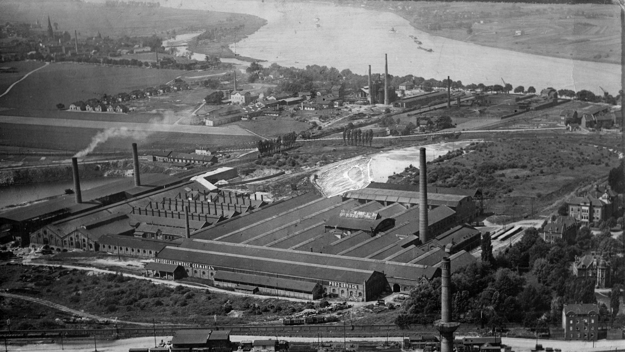 Das Schwarz-Weiß-Luftbild zeigt das Werksgelände der Spiegelglaswerke Germania in Porz um 1900 mit Industriehallen und Schornsteinen, im Hintergrund ist der Rhein zu sehen.