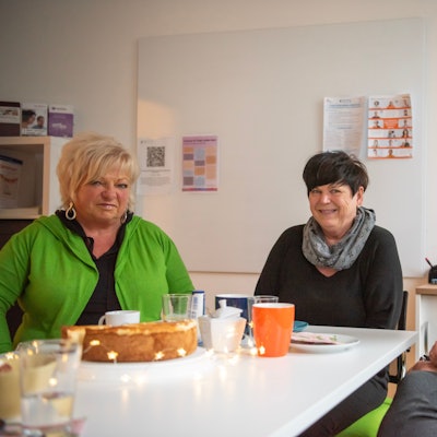 Die drei Frauen sitzen an einem Tisch in einem Büroraum. Auf dem Tisch stehen ein Käsekuchen und Kaffeegeschirr, daneben liegt eine Sternen-Lichterkette.