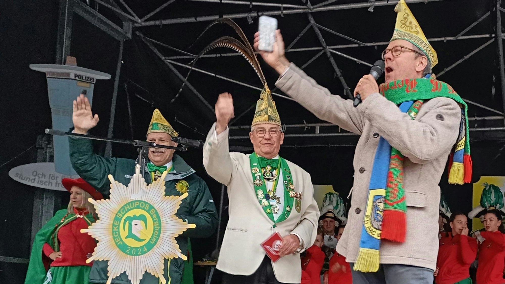 Drei Männer mit Narrenkappen stehen auf einer Bühne.