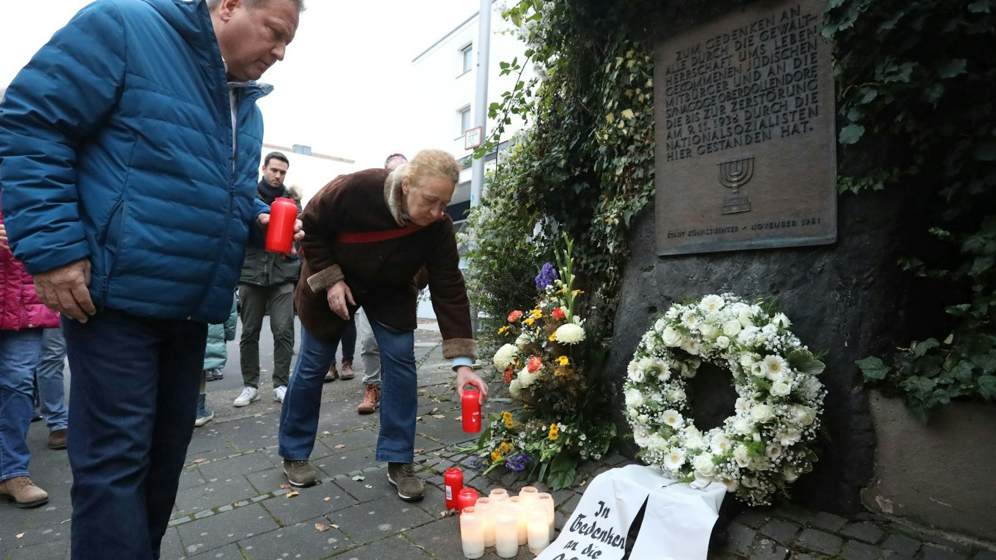 Kränze, Blumen und Kerzen vor einer Bronzetafel.